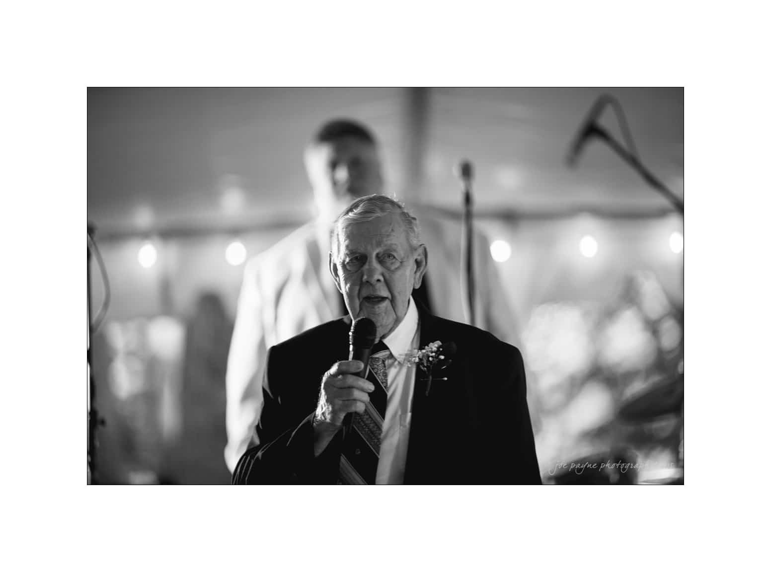NC wedding photography - rachel & joe Black and white photo of an older man in a suit speaking into a microphone. The background is out of focus, featuring soft lights and a partially visible person standing behind him.