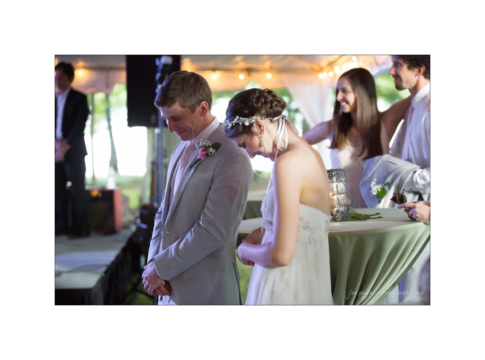 NC wedding photography - rachel & joe A bride and groom bow their heads in prayer during a wedding ceremony. They stand close together, surrounded by guests in a softly lit venue with decorations. A bridesmaid and groomsman are visible in the background.