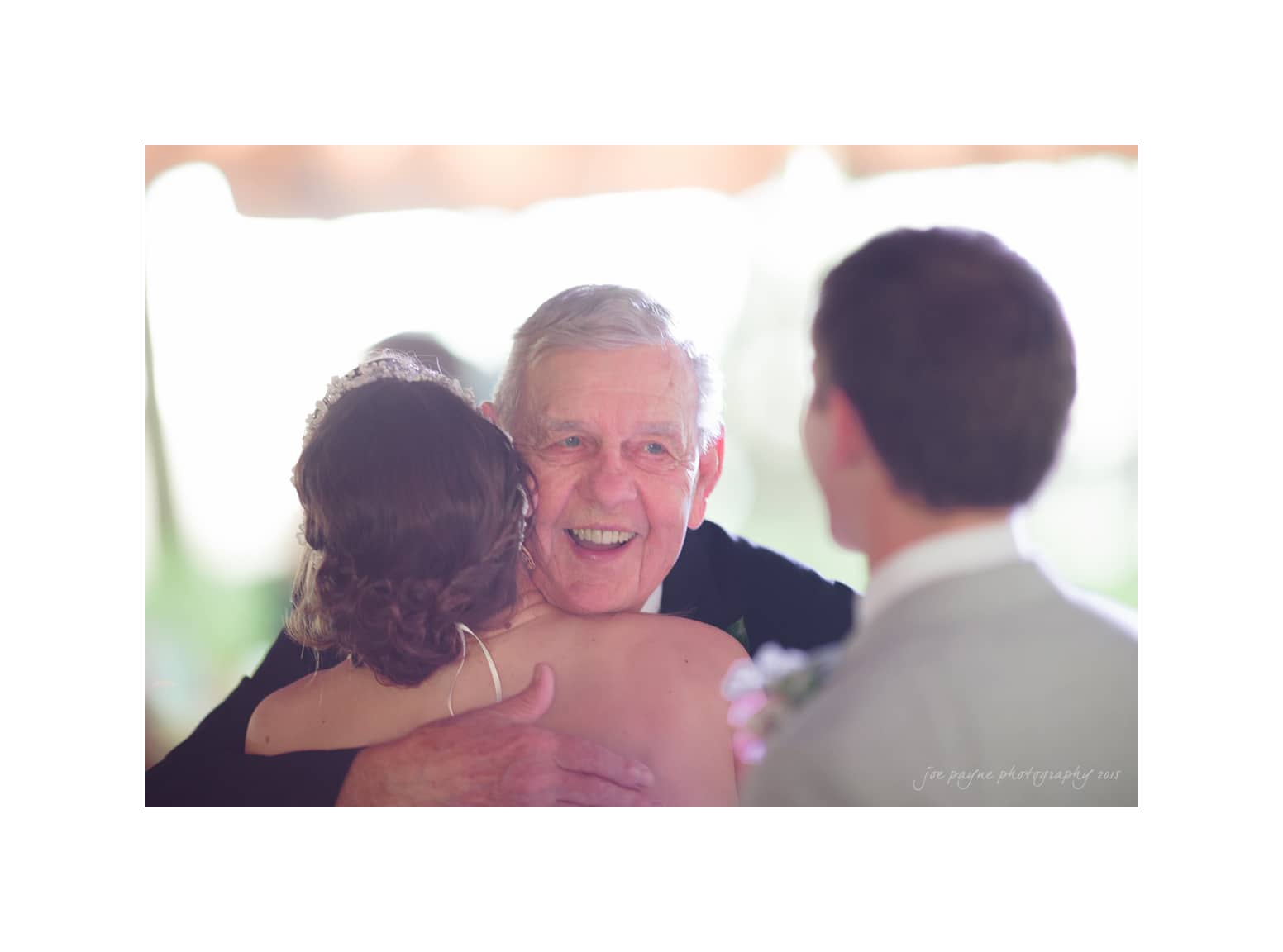 NC wedding photography - rachel & joe An elderly man smiles warmly while hugging a woman in a bridal gown, as a man in a suit looks on. The setting suggests a wedding celebration with soft lighting and a joyful atmosphere.