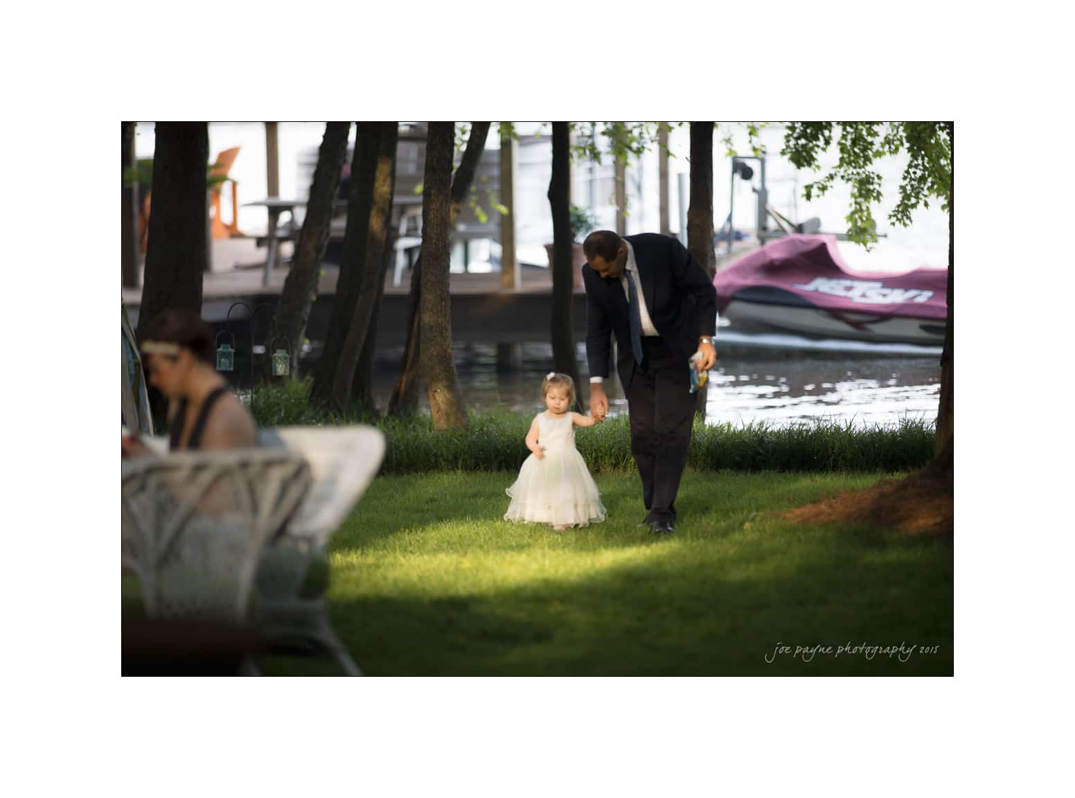 NC wedding photography - rachel & joe A man in formal attire walks hand-in-hand with a young girl in a white dress across a grassy area. Trees and boats are visible in the background. The scene is outdoors and appears serene.