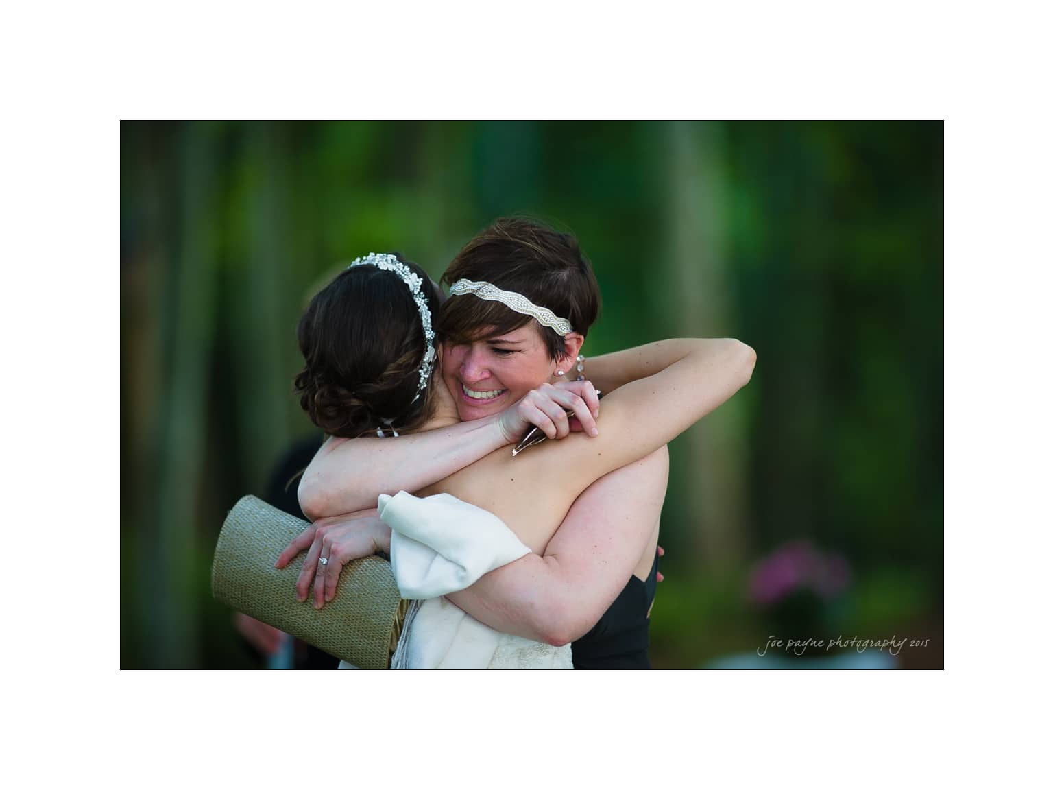 NC wedding photography - rachel & joe Two women embracing warmly, both smiling with eyes closed. One woman wears a white dress with a headpiece, the other in a black dress with a headband. Background shows blurred greenery.