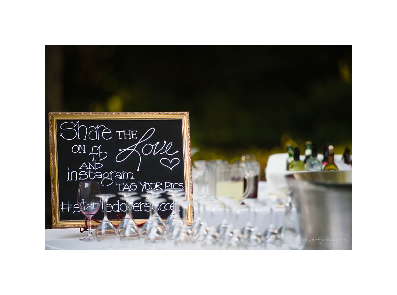 NC wedding photography - rachel & joe A chalkboard sign on a table displays the message urging guests to share the love on social media, highlighting a hashtag. The table is set with wine glasses, bottles, and a large ice bucket, against a blurred outdoor background.