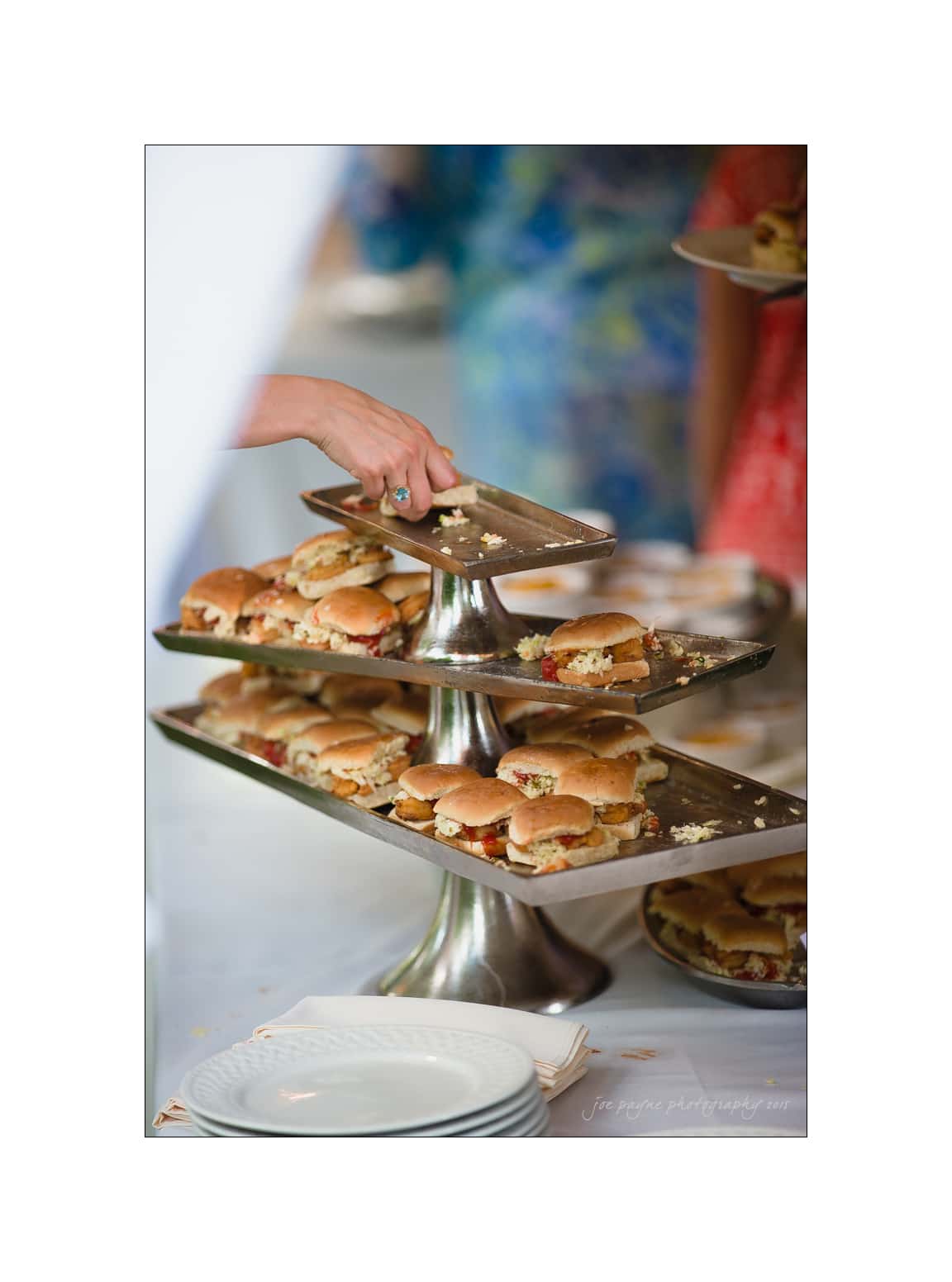 NC wedding photography - rachel & joe A hand reaching for a small sandwich on a tiered serving tray filled with sliders. The trays are stacked on a table covered with a white tablecloth, and there are empty plates nearby, in a buffet setting.