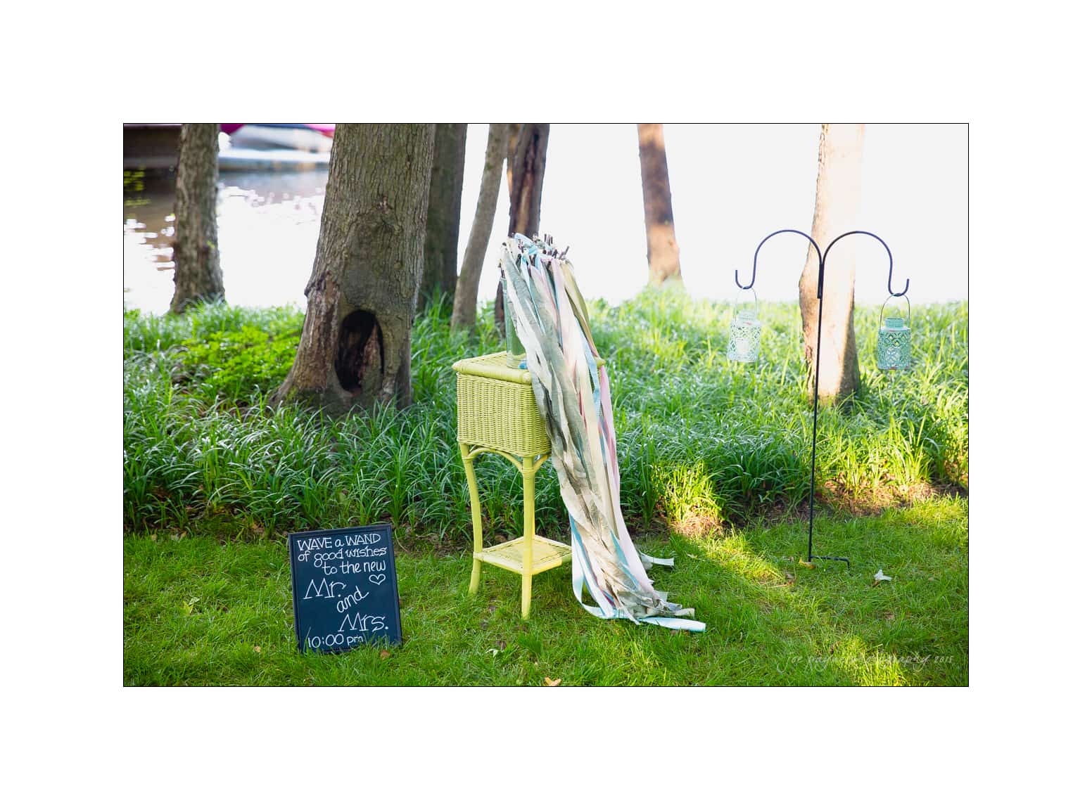 NC wedding photography - rachel & joe A grassy outdoor setting features a yellow wicker stand with rolled blankets, a chalkboard sign with Take a blanket for the boat ride, and two lanterns on metal hooks. Trees and a body of water are in the background.