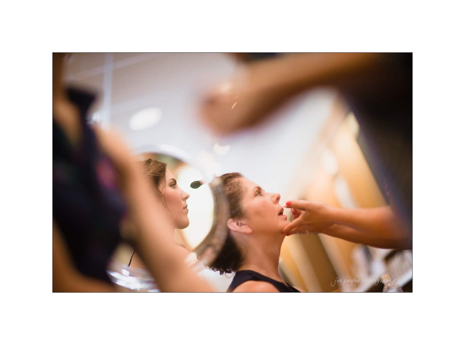 NC wedding photography - rachel & joe A person is having makeup applied while sitting in front of a mirror. Another person oversees the process. The scene is softly focused, emphasizing the act of applying lipstick and the reflection in the mirror.