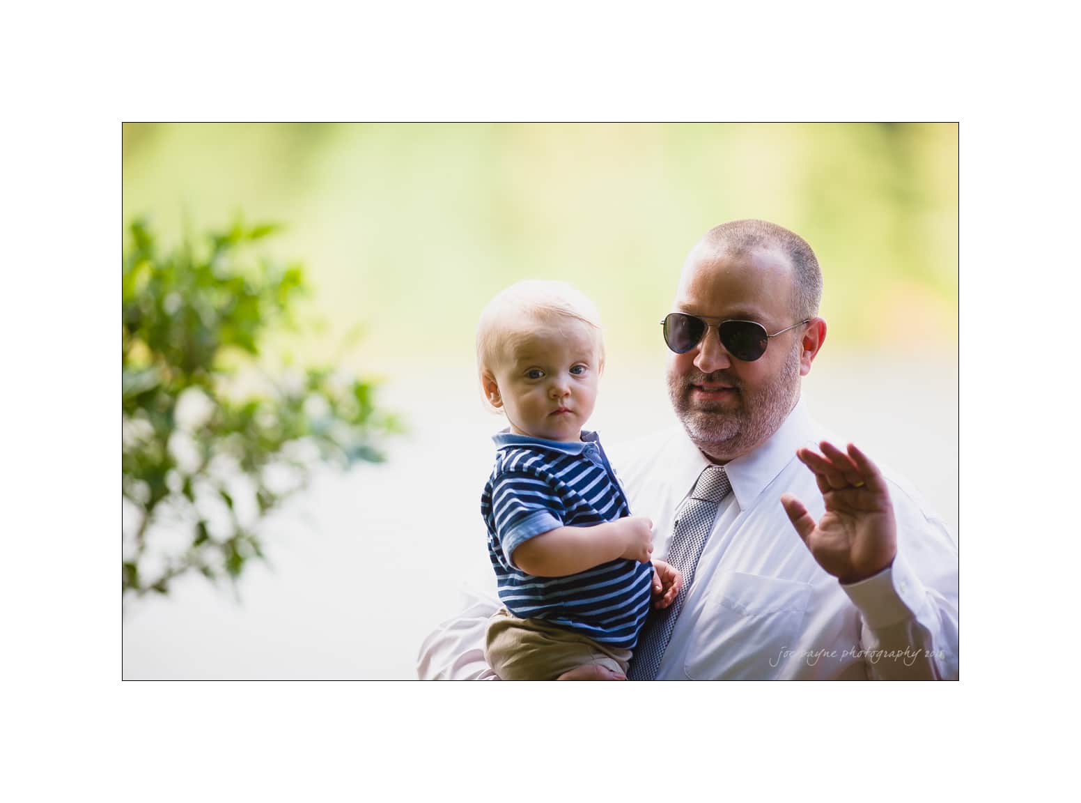 NC wedding photography - rachel & joe A man wearing sunglasses and a tie is holding a baby in his arms outdoors. The baby is wearing a blue striped shirt. They are both looking at the camera, and the background is green and blurred.