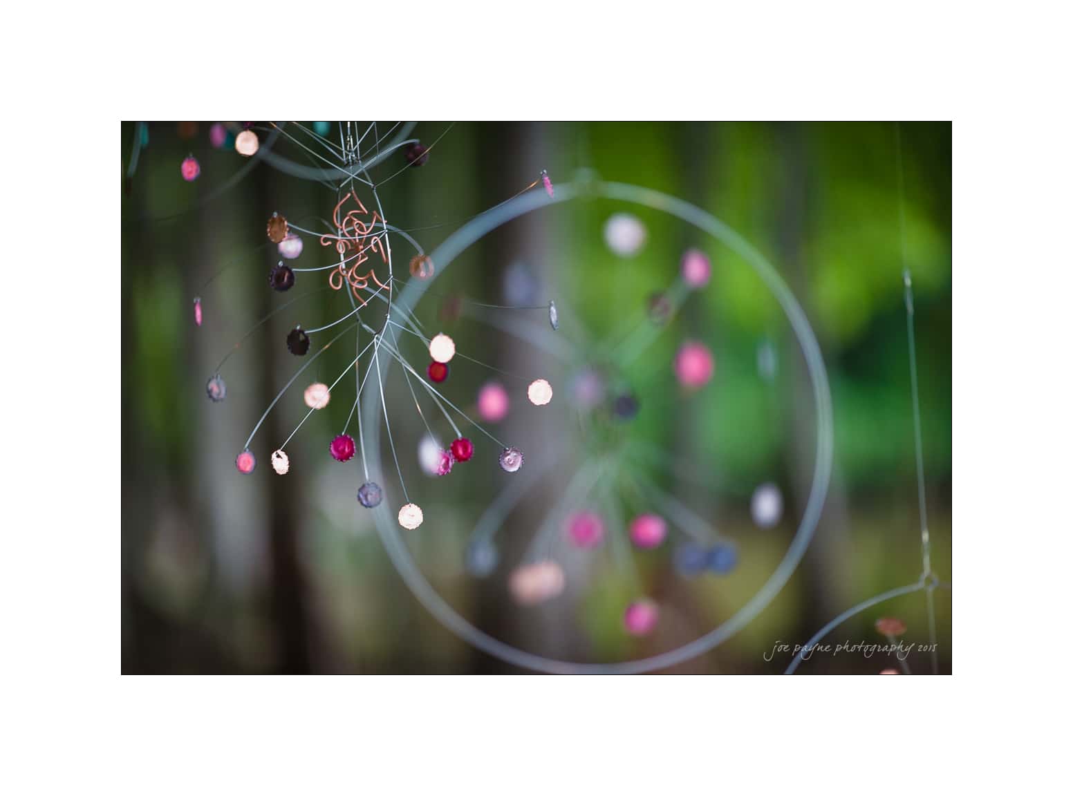 NC wedding photography - rachel & joe A delicate mobile hangs from thin wires, adorned with small, colorful discs in shades of red, pink, and black, set against a blurred green background.