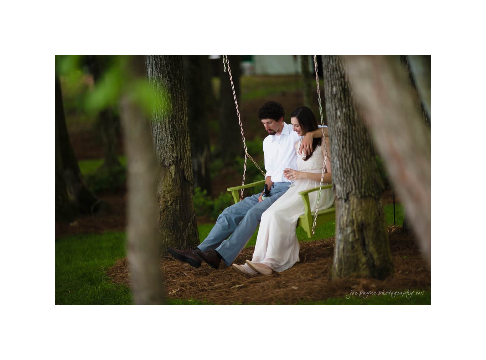 NC wedding photography - rachel & joe A couple sits closely on a swing hanging from trees, surrounded by greenery. The man wears a white shirt and blue pants, while the woman is in a long white dress. They appear relaxed and content, enjoying the peaceful outdoor setting.