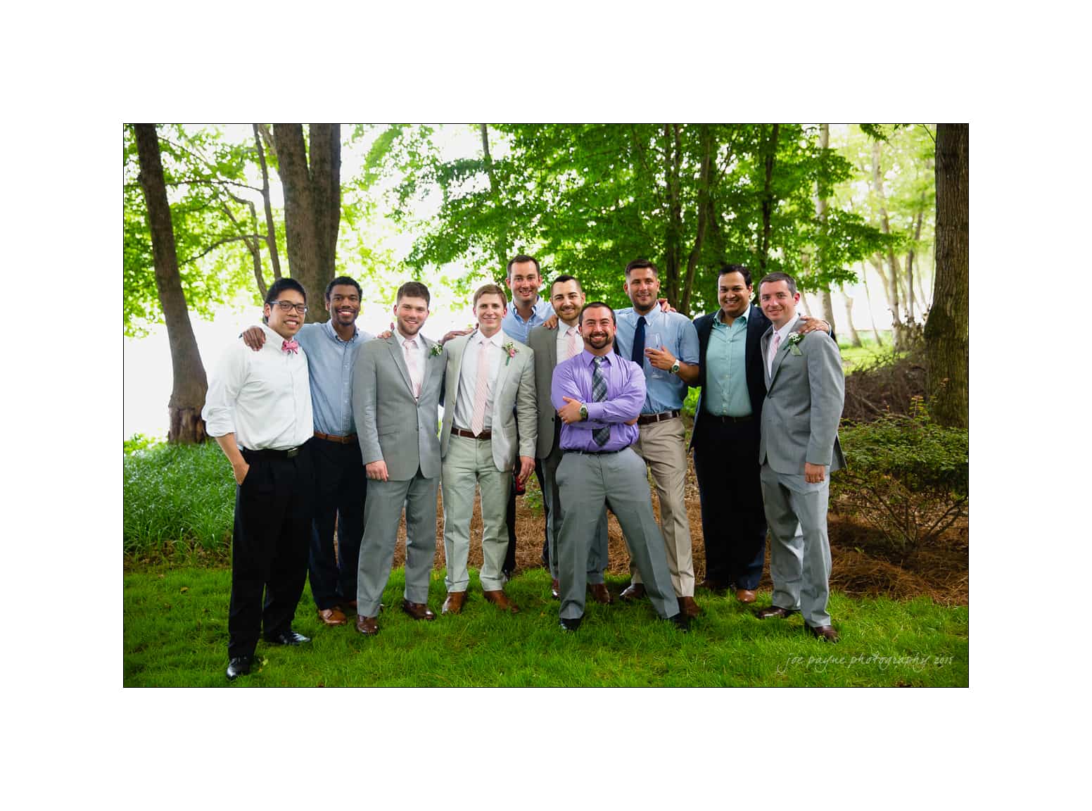 NC wedding photography - rachel & joe A group of eleven men dressed in formal attire stand together outdoors on a lawn, surrounded by trees. They are smiling and have their arms around each others shoulders.