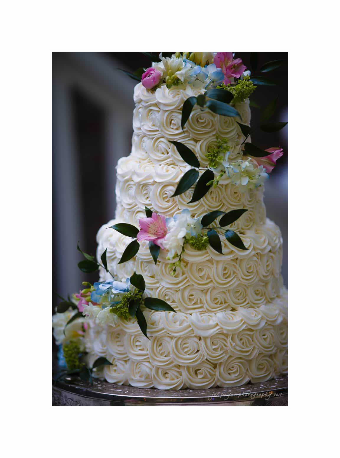 NC wedding photography - rachel & joe A three-tiered wedding cake adorned with intricate white frosting patterns resembling roses. Decorated with pink, blue, and green flowers and leaves, the cake sits elegantly on a silver stand.