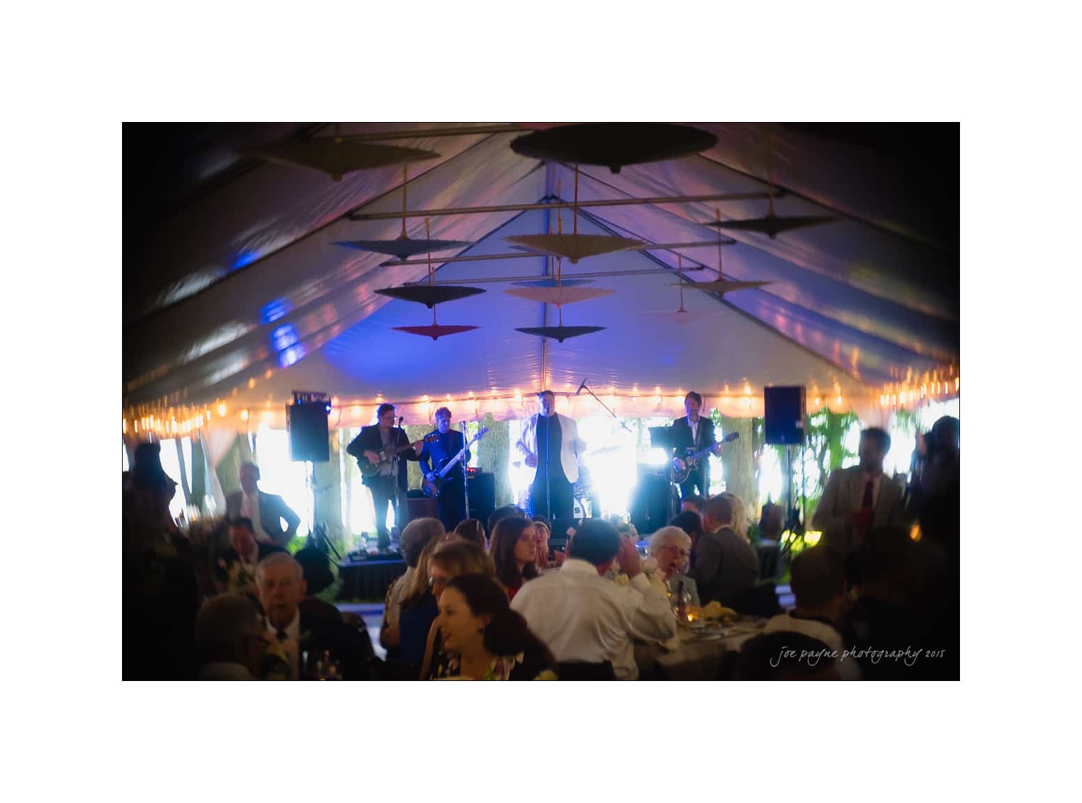 NC wedding photography - rachel & joe A live band performs on a stage at an event under a large tent with string lights. People seated at tables in the foreground are enjoying the music, creating a lively and festive atmosphere.