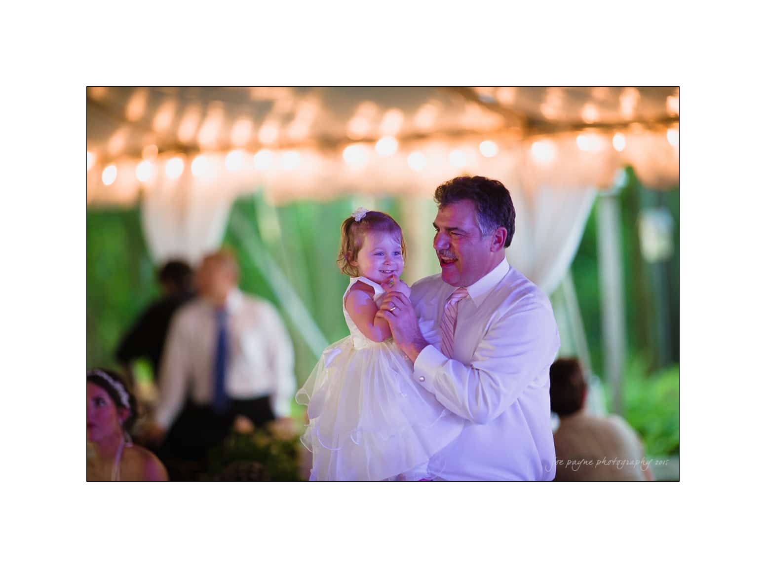 NC wedding photography - rachel & joe A man is holding a young child dressed in a white dress at an outdoor event. They are both smiling. The background is softly lit with string lights and blurred greenery. It appears to be a joyful and festive occasion.