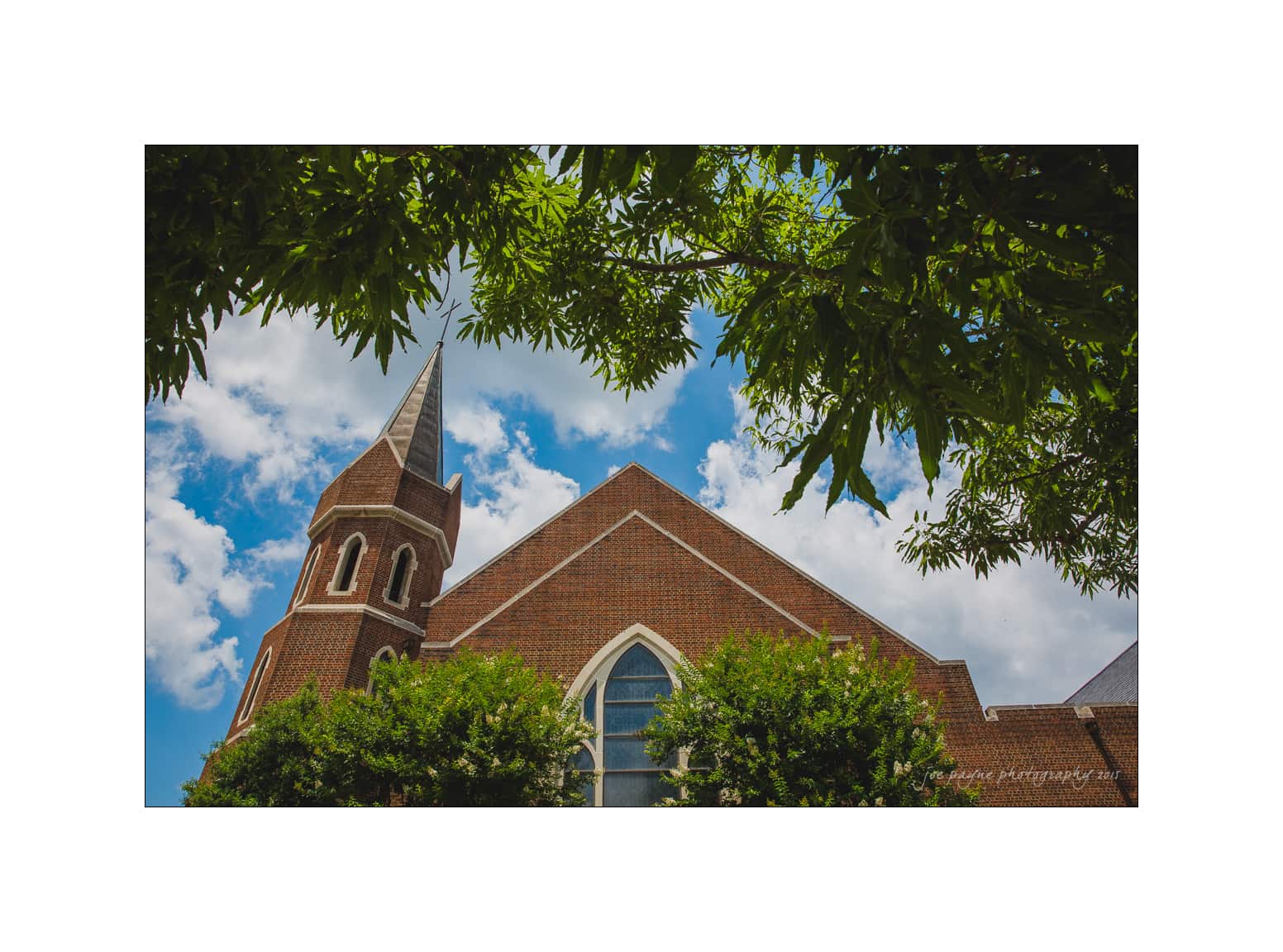 NC wedding photography - rachel & joe A red brick church with a tall steeple and arched windows is framed by green leafy branches. The sky is blue with scattered clouds.