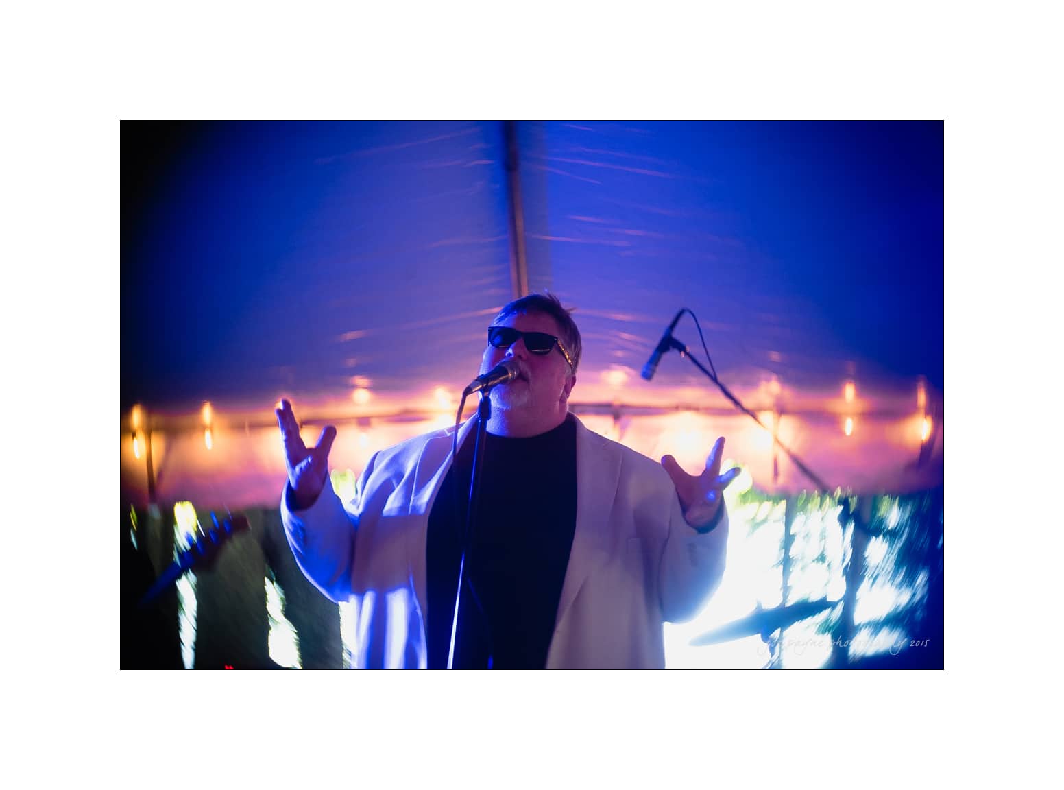 NC wedding photography - rachel & joe A person wearing sunglasses and a light-colored jacket performs energetically on stage under purple and blue lights. They hold a microphone, with instruments visible in the background.