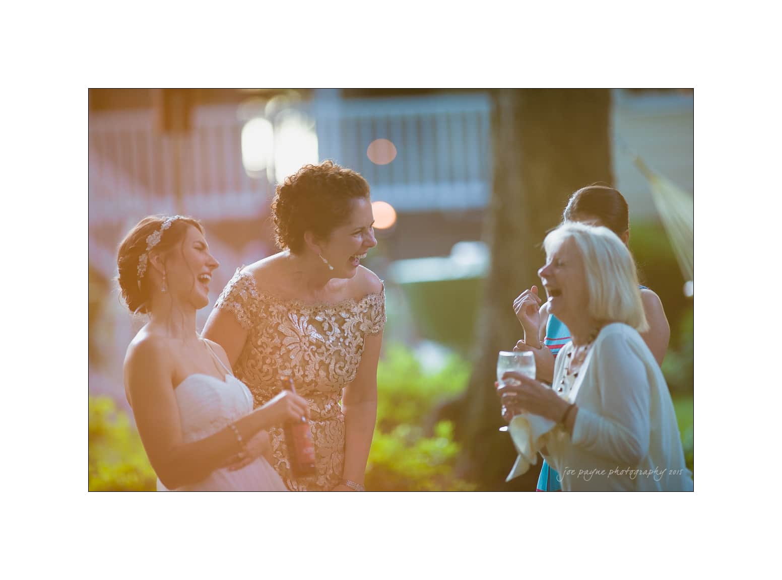 NC wedding photography - rachel & joe Three women laughing and enjoying a conversation outdoors. One is a bride in a white dress, another wears an elegant lace dress, and the third is holding a drink, wearing a light sweater. The scene is warmly lit, suggesting a joyful event.