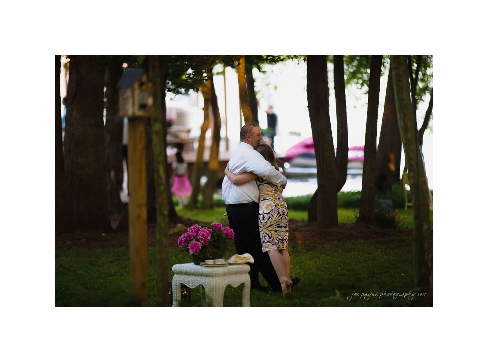 NC wedding photography - rachel & joe A couple embraces while dancing in a wooded outdoor setting. The man wears a white shirt and dark pants, and the woman is in a patterned dress. A small table with pink flowers is in the foreground, and trees are visible in the background.