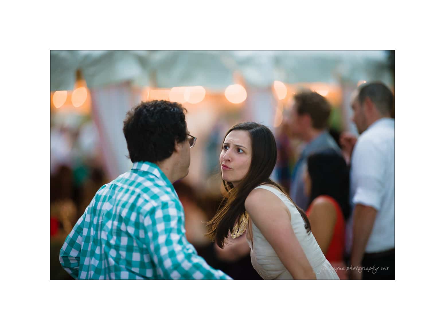 NC wedding photography - rachel & joe A woman in a white dress and a man in a checkered shirt engage in an intense conversation at an outdoor event. Blurred attendees and string lights are visible in the background.