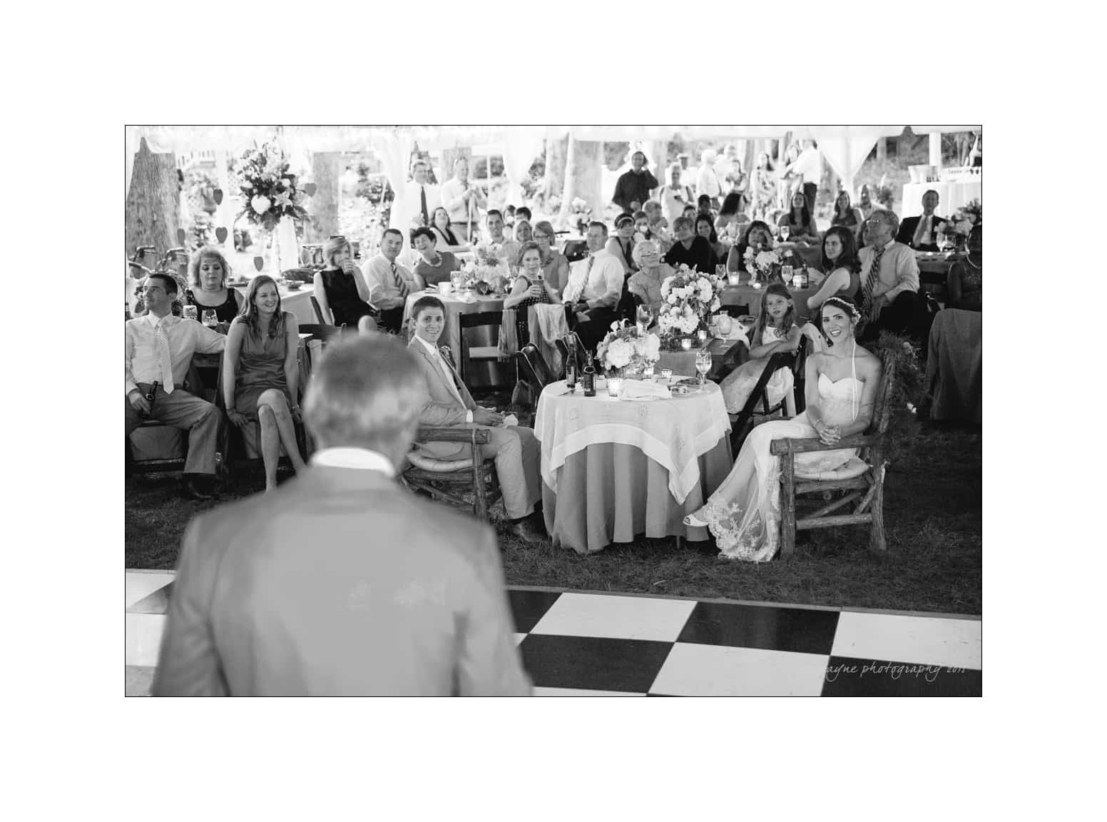 NC wedding photography - rachel & joe A black and white photo of a wedding reception with guests seated at tables, listening attentively to a person speaking. The bride and groom are seated together, smiling, near a checkered dance floor under a tent.