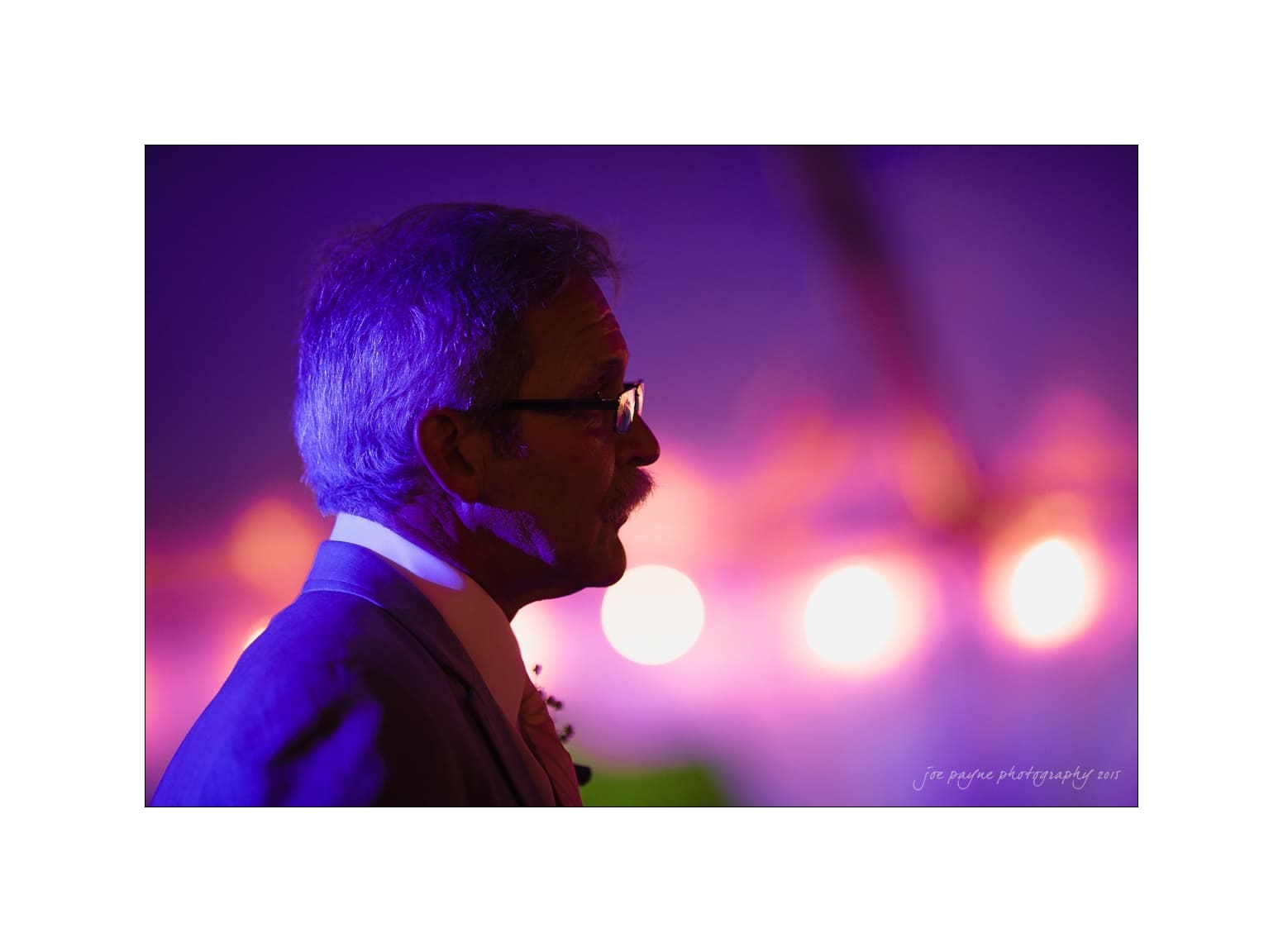 NC wedding photography - rachel & joe Profile of a man silhouetted against a vibrant purple and pink background, wearing glasses and a suit. The background is softly lit, creating a colorful ambiance.