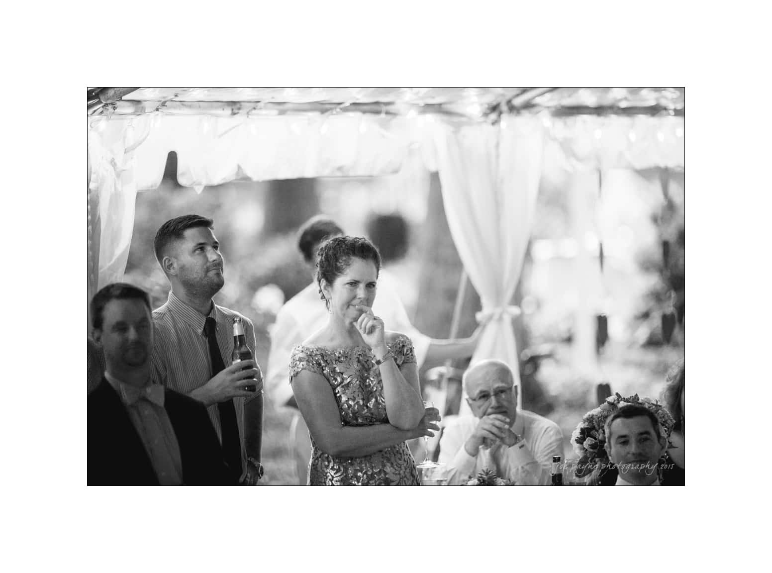 NC wedding photography - rachel & joe A black and white photo of people at a formal gathering. A woman in a floral dress is smiling, a man next to her holds a beer. Other attendees are seated in the background under a canopy with draped curtains.