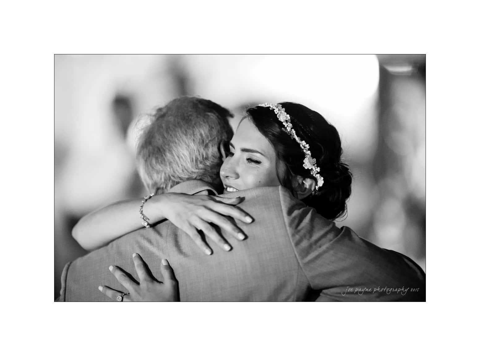 NC wedding photography - rachel & joe A black and white photo of a woman wearing a floral headband, embracing an older man in a suit. They are both smiling, sharing a warm hug, likely during a special occasion.