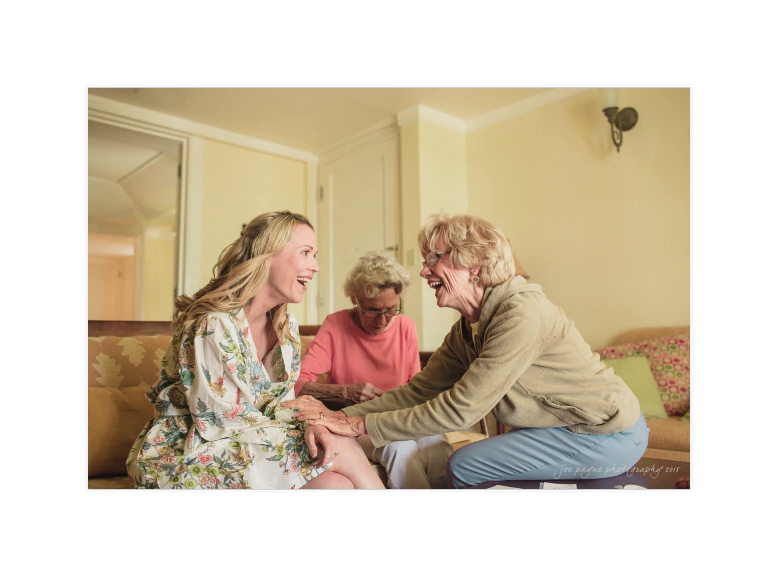 A joyful moment captured as three women gather in a cozy room. One woman in a floral robe sits on a sofa, smiling, while another leans toward her, holding her hands warmly. A third woman with white hair observes, smiling gently.