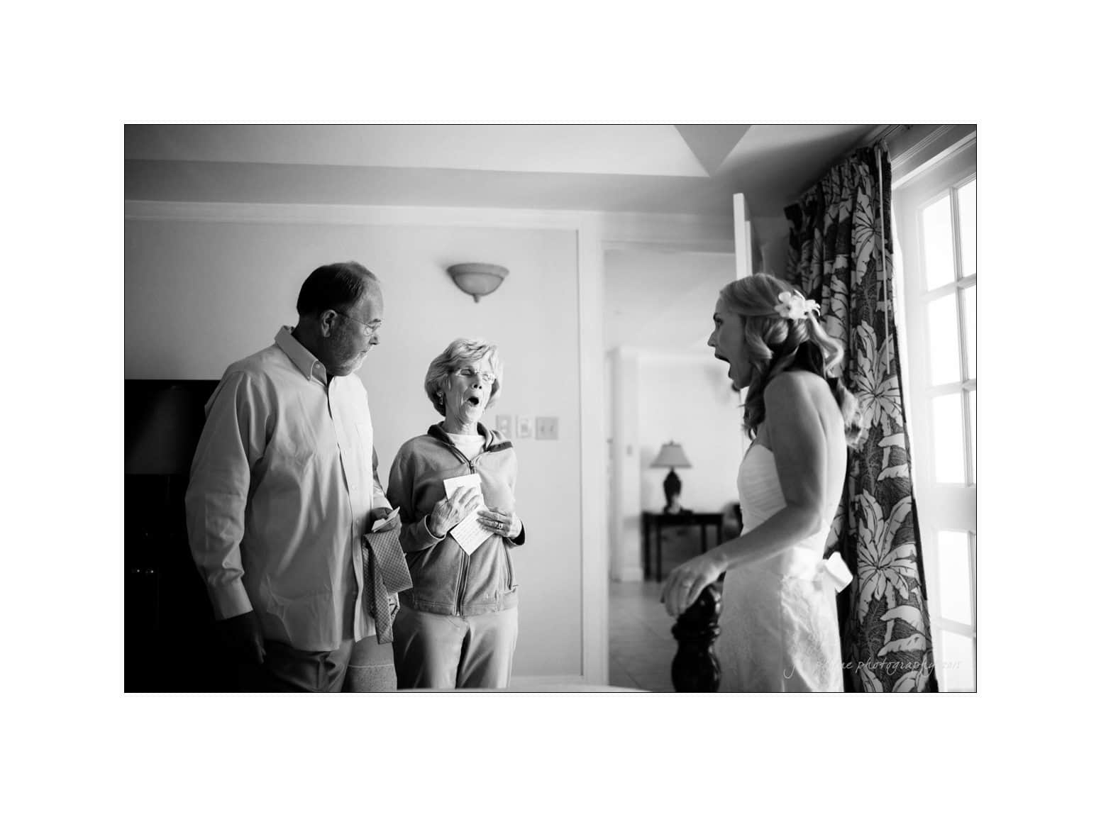 A bride stands in a room, wearing a white gown, looking surprised. She faces two people, a man and woman, who are also gazing at her. The room is softly lit and has a window with patterned curtains in the background.
