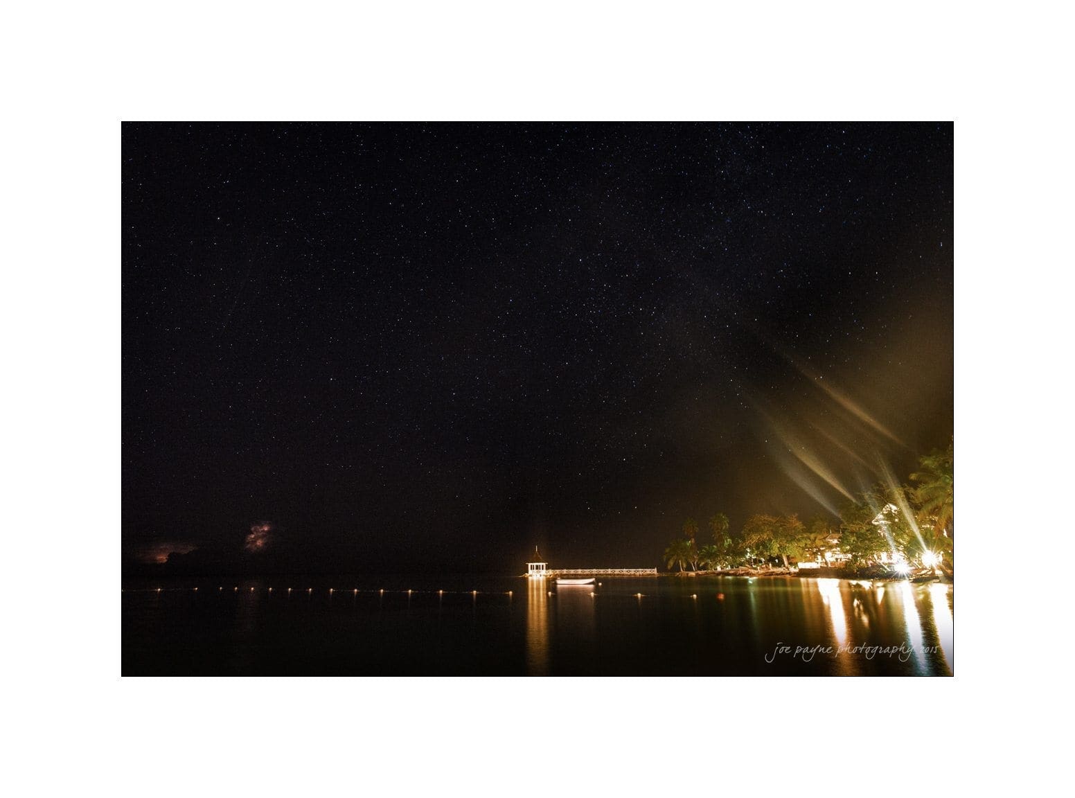 A serene night scene with a star-filled sky above a calm body of water. A pier extends into the water, illuminated by lights on the right, with trees nearby. Light reflections shimmer on the waters surface.