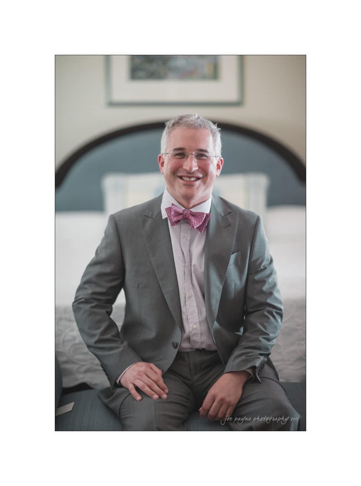 A man in a gray suit with a pink bow tie sits on a bed. He is smiling and wearing glasses. The background features a blurred headboard and framed artwork on the wall.