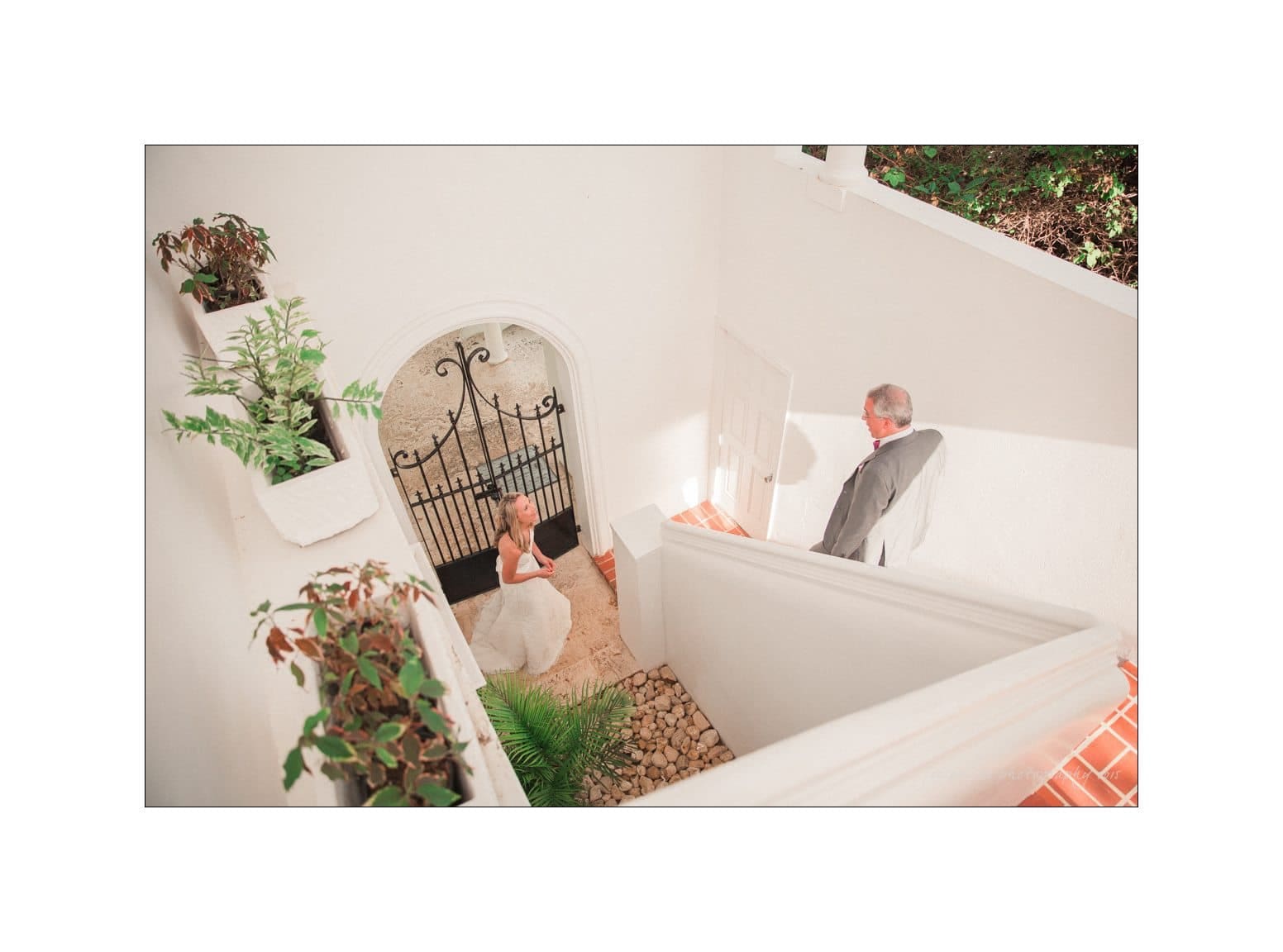 A bride and an older man stand in a white, multi-level courtyard with potted plants. The bride, in her wedding dress, looks up at the man, possibly her father, who is descending the stairs. The scene is lit by natural sunlight.