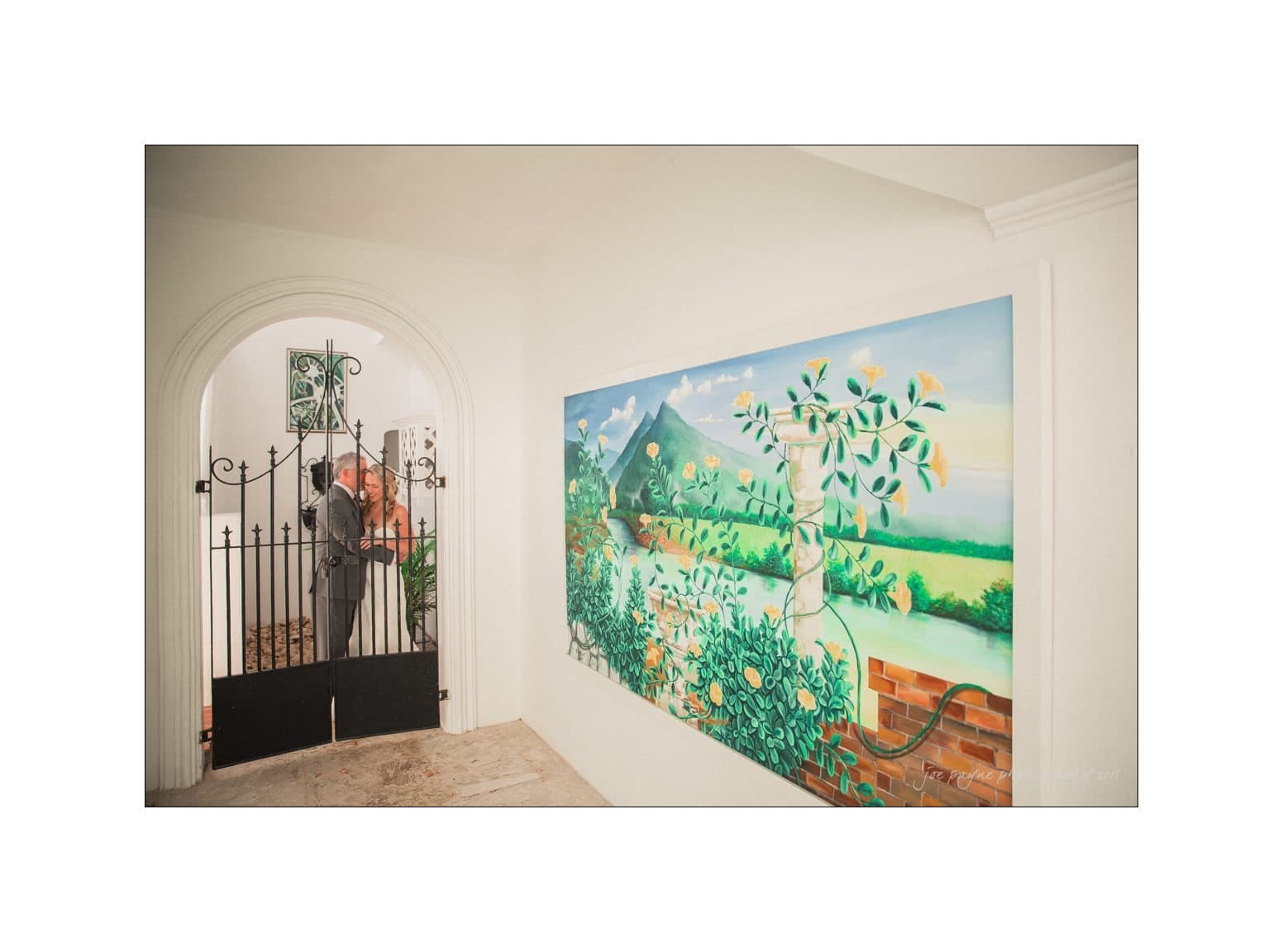 A person opens a black wrought-iron gate in a white hallway. A large painting on the right depicts a vibrant garden with trees and a distant mountain.