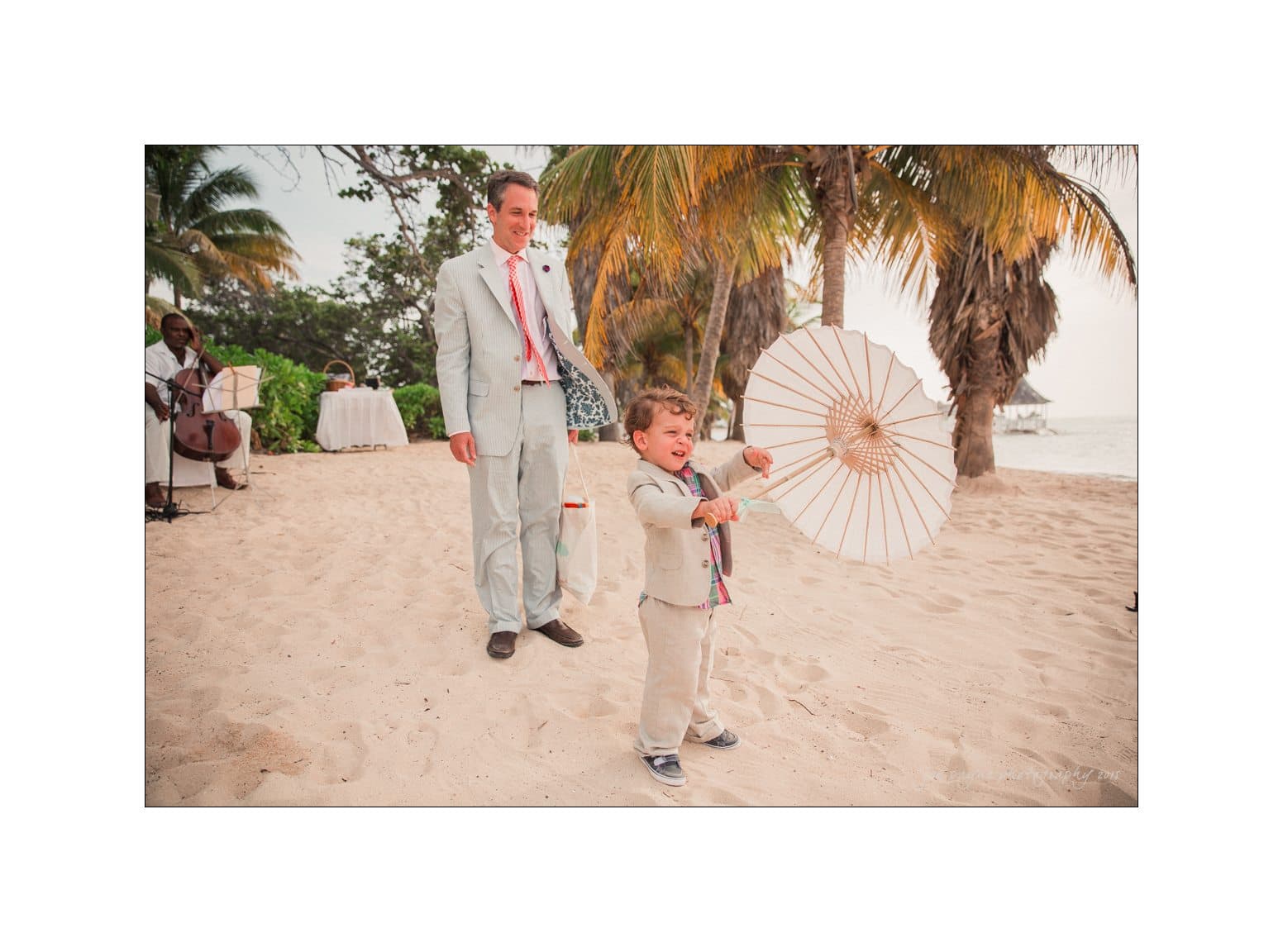A joyful child in a light gray suit holds a white parasol on a sandy beach, with a smiling adult in a matching suit nearby. Tropical trees and the ocean are in the background.