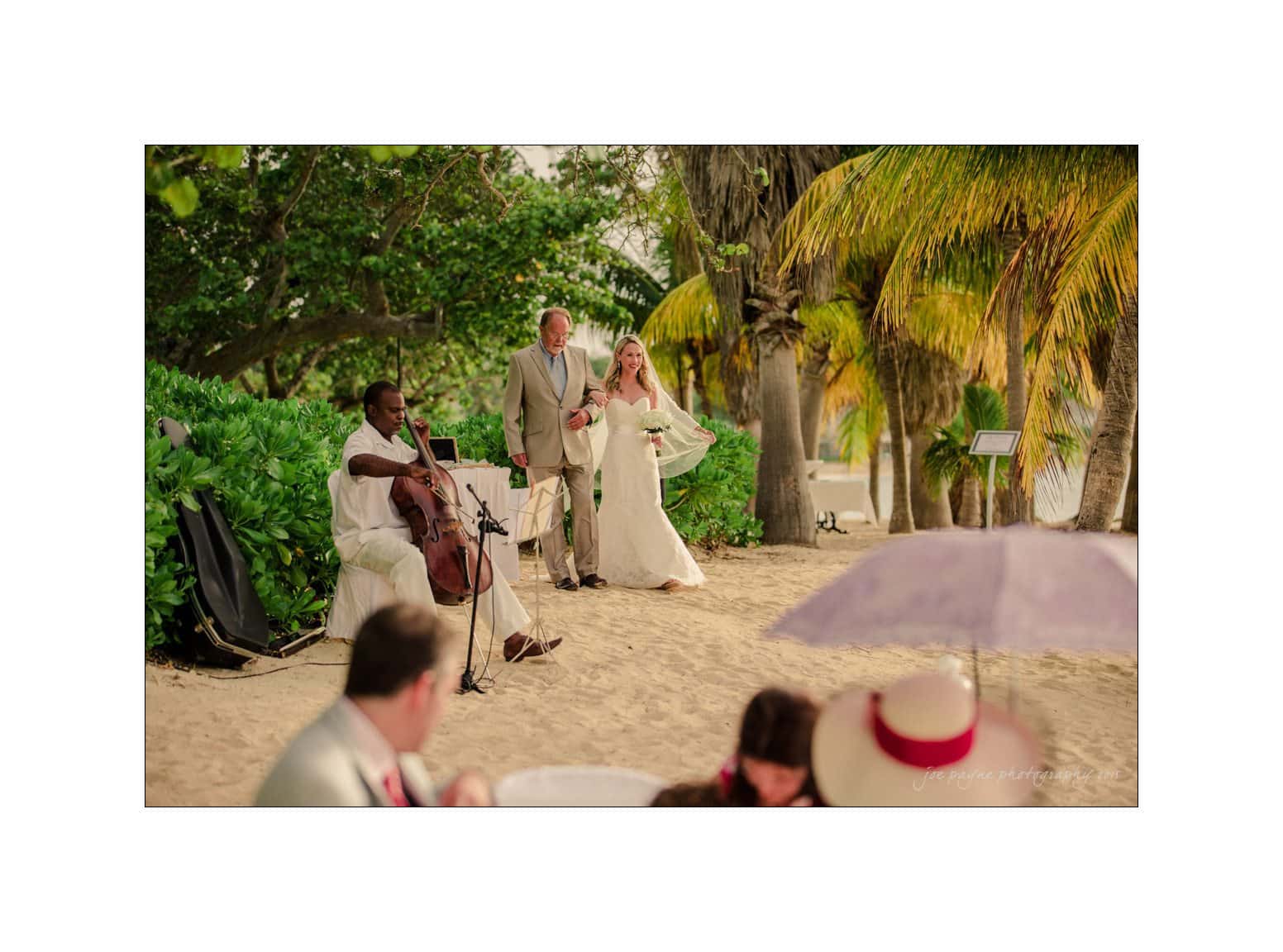 A bride walks joyfully down a sandy beach aisle, arm in arm with a suited man, as a cellist plays nearby. Lush palm trees and greenery surround them, creating a tropical backdrop, while guests sit under umbrellas in the foreground.