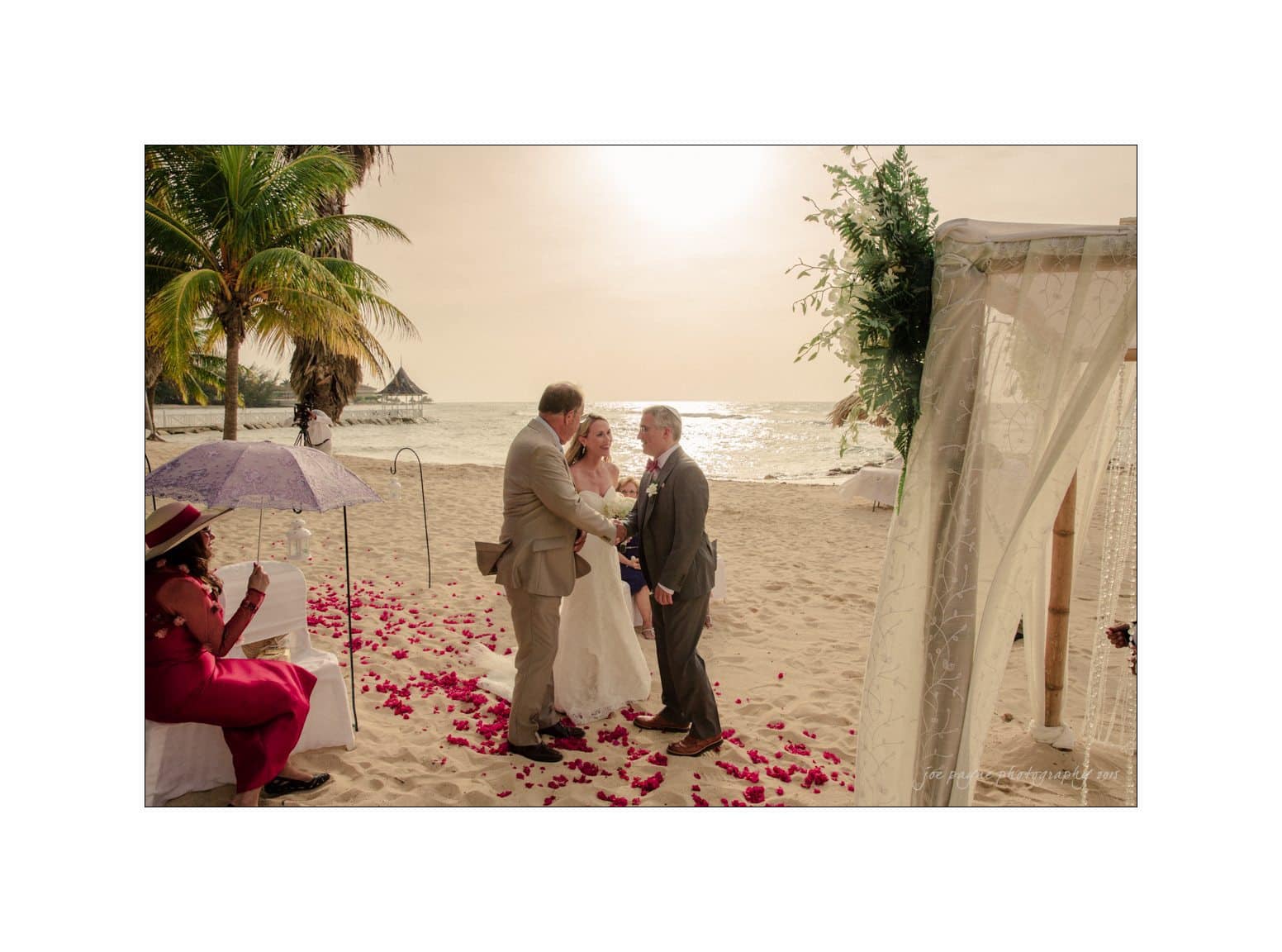 A bride and groom stand together on a beach for their wedding ceremony. The officiant is speaking to them. Theres an arch decorated with fabric and flowers, and red petals scattered on the sand. Guests are seated, and palm trees frame the scene.