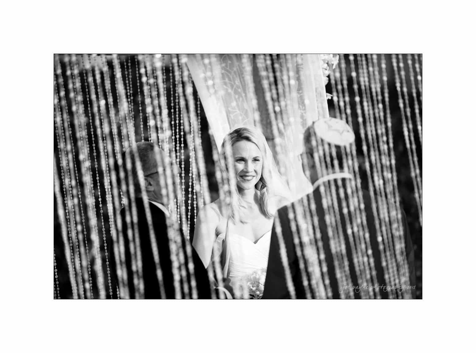 A black-and-white photo of a bride smiling during a wedding ceremony. She stands opposite two people, partially obscured by hanging strings or beads.