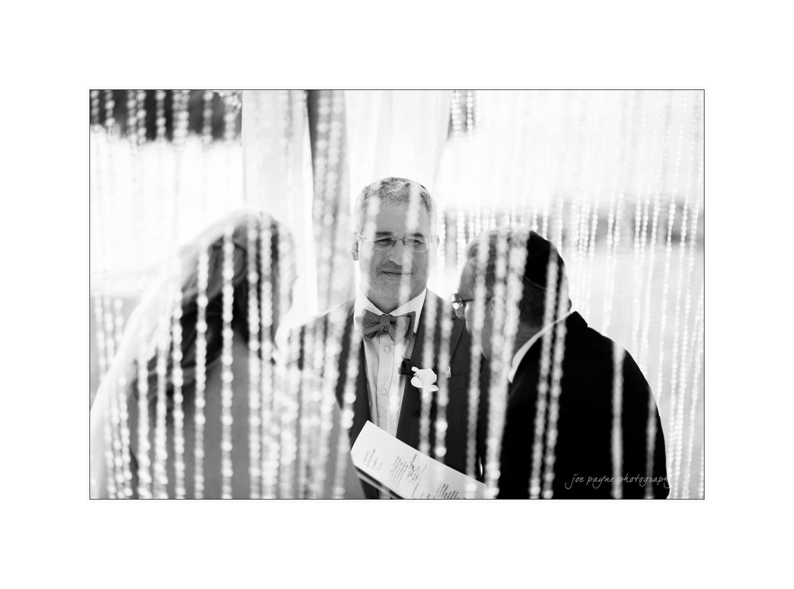 A black and white photo of a wedding ceremony, seen through hanging beads. Two people face the officiant, who is holding a booklet. The setting appears intimate and elegant.