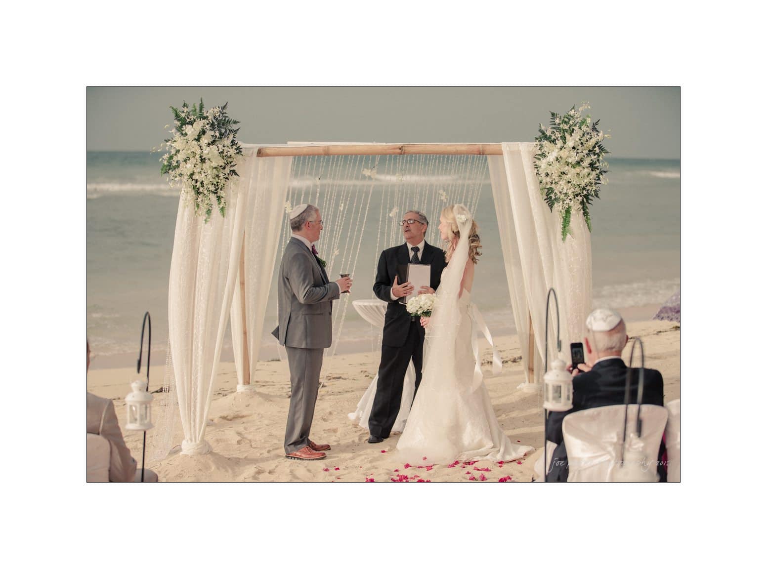 A couple in wedding attire stands under a floral chuppah on a sandy beach, exchanging vows with a rabbi. Guests are seated nearby, and the ocean is in the background.