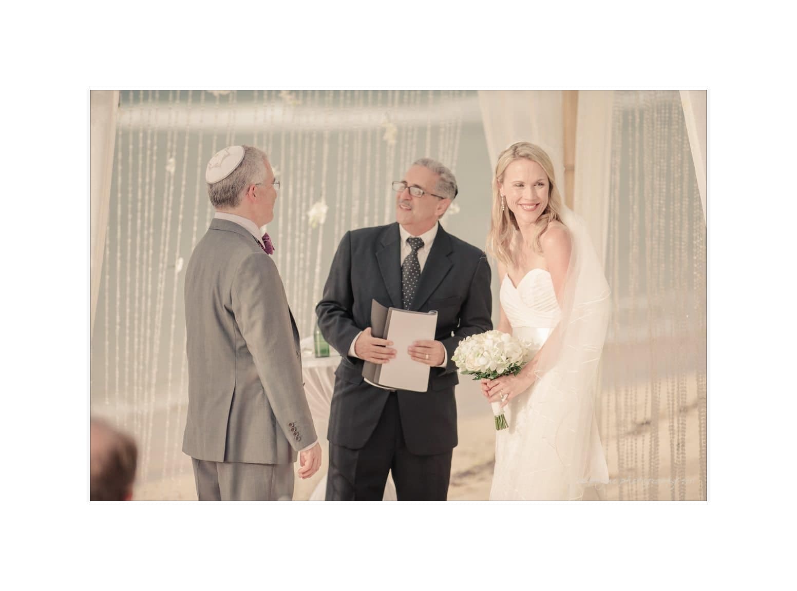 A bride in a white dress holds a bouquet while standing next to a groom in a gray suit. They are under a canopy, with an officiant in a suit smiling at them. The setting appears to be outdoors on a beach.