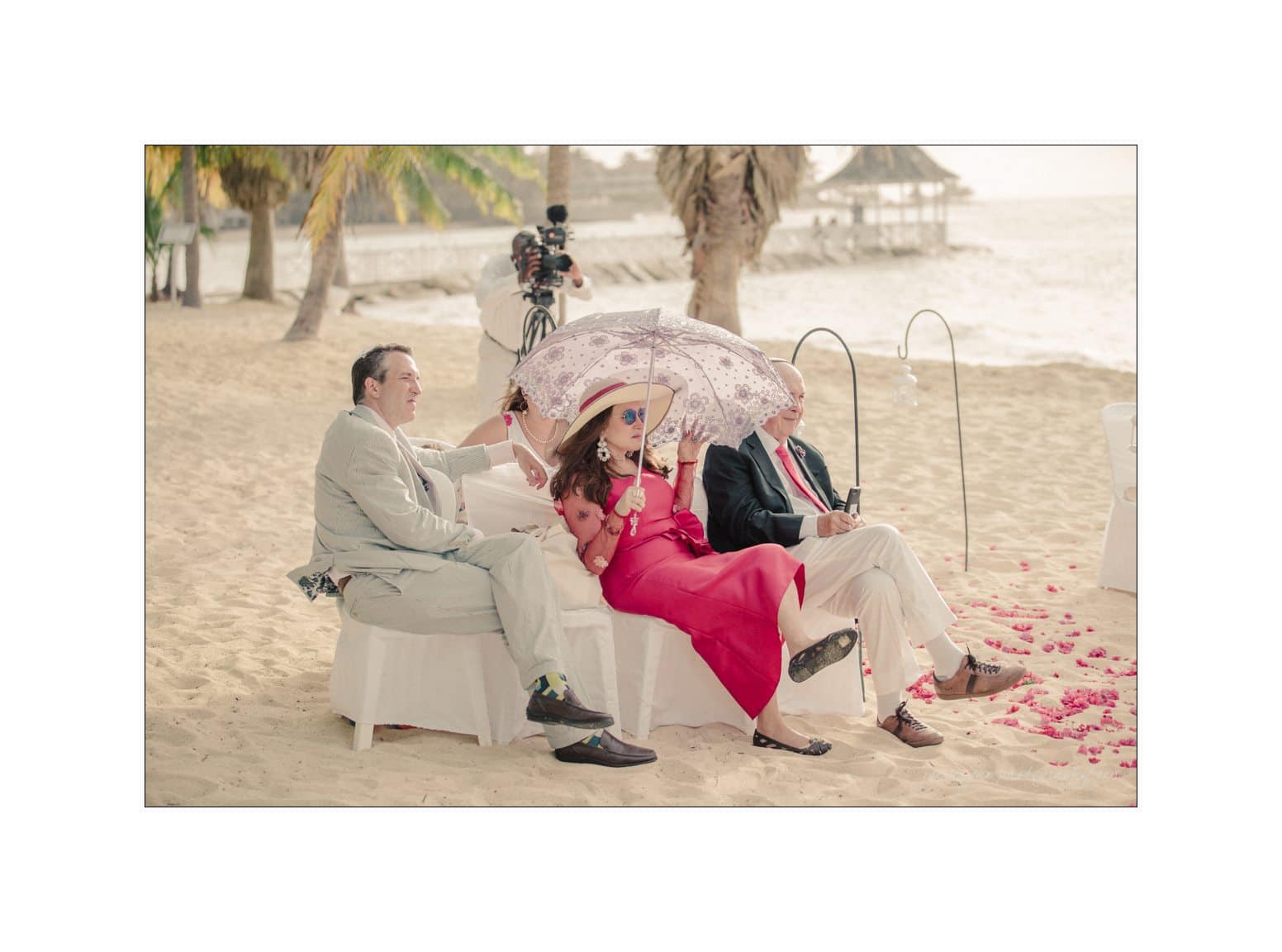 Three people sit on a white couch at a beach, with palm trees and the sea in the background. The woman in the middle, wearing a red dress, holds a floral umbrella. Pink flower petals are scattered on the sand beside them.