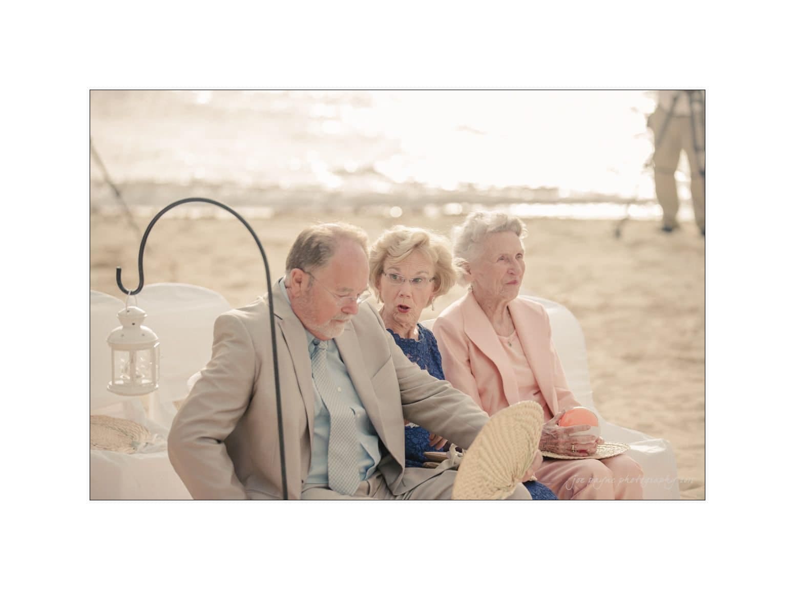 Three elderly people sit on a beach. A man in a beige suit adjusts his shoe while two women, one in a blue dress and the other in pink, watch. A lantern hangs nearby, and the background shows a sandy shore.