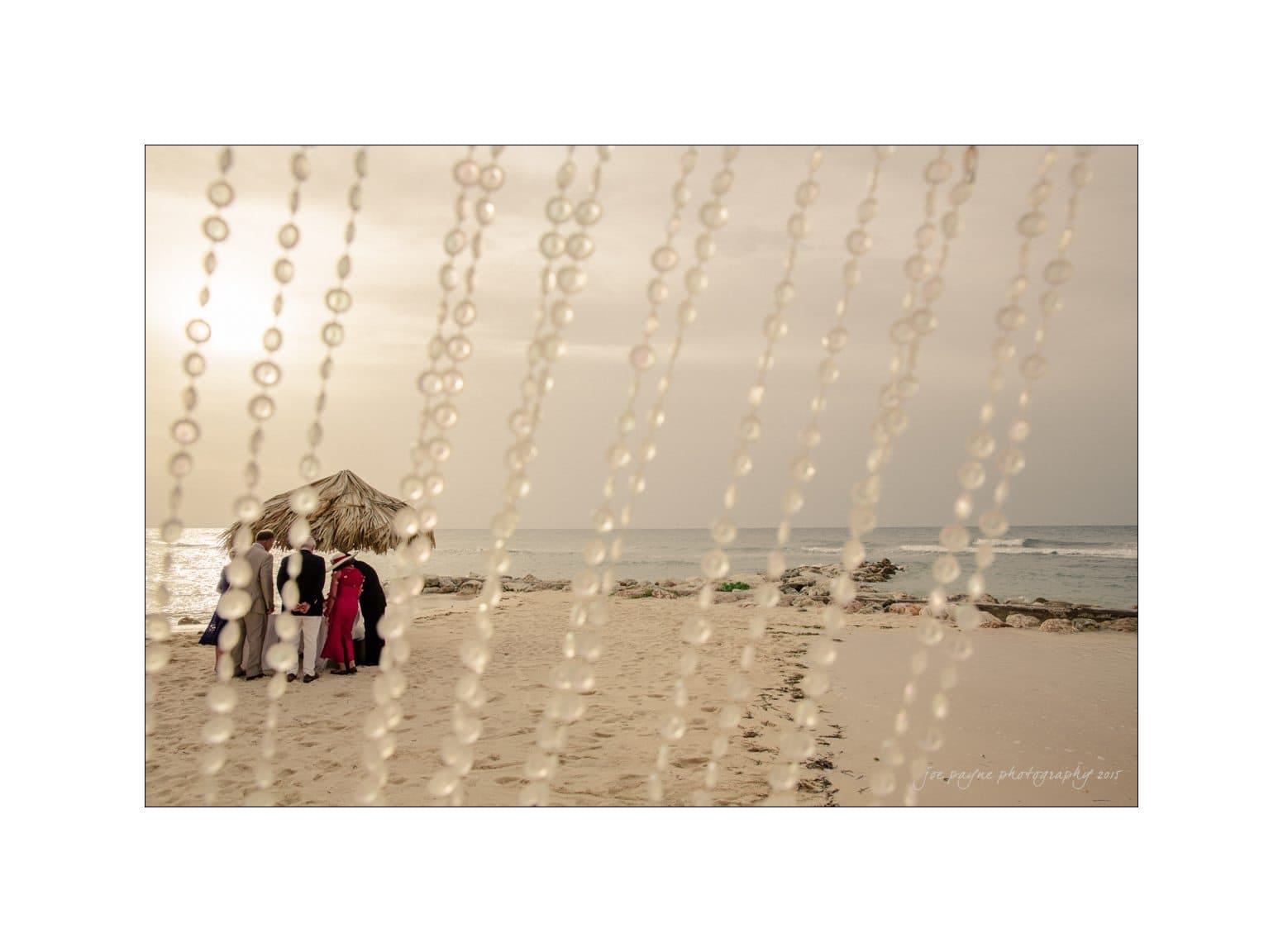 A beach wedding scene viewed through hanging beads. A group of people stand near a small thatched-roof structure on sandy shores, with the ocean in the background. The image has a soft, warm tone.
