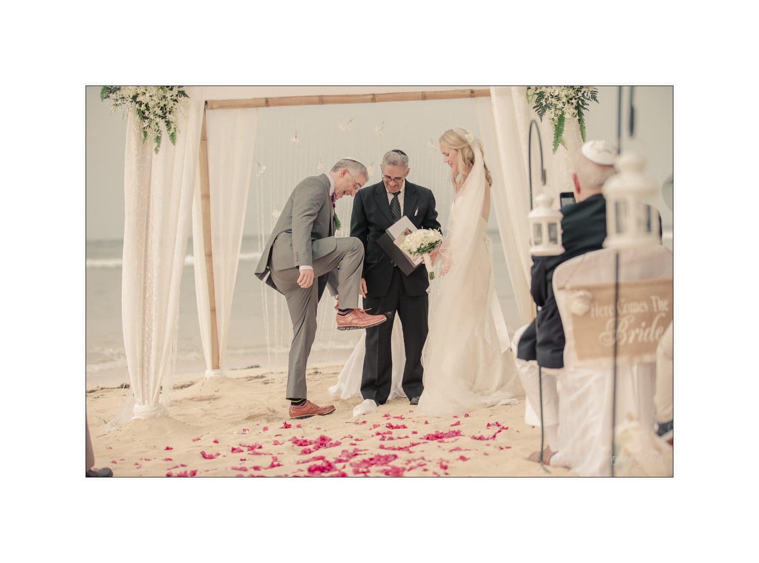 A beach wedding scene with a couple under a decorated canopy. The groom playfully lifts his leg as the officiant holds a book. Rose petals are scattered on the sand, and a sign partially visible reads Here comes the Bride.
