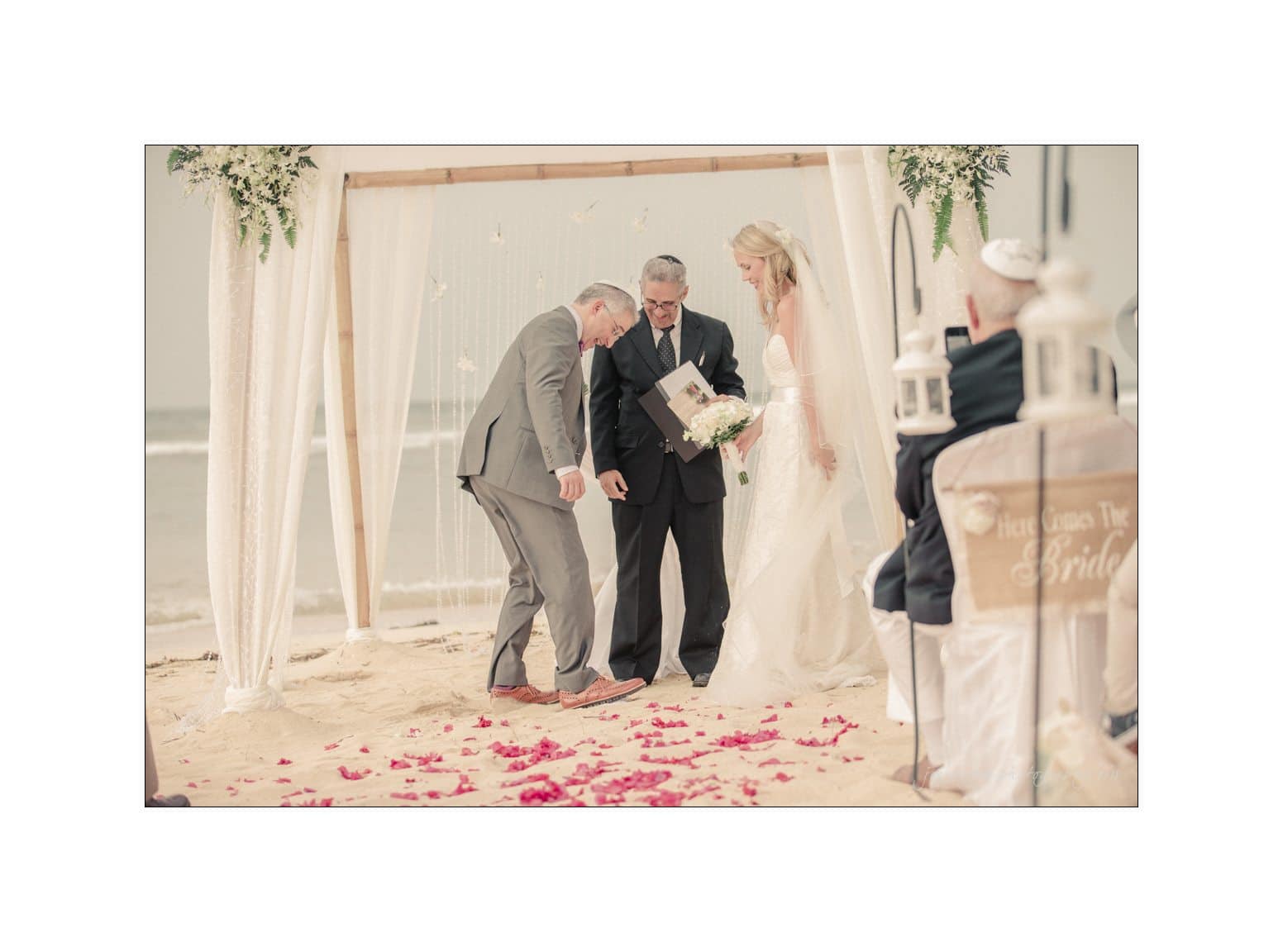 A couple stands under a wooden canopy on a sandy beach during a wedding ceremony. The officiant holds a book, red petals scattered on the sand. Guests are seated nearby, one wearing a kippah. The ocean is visible in the background.