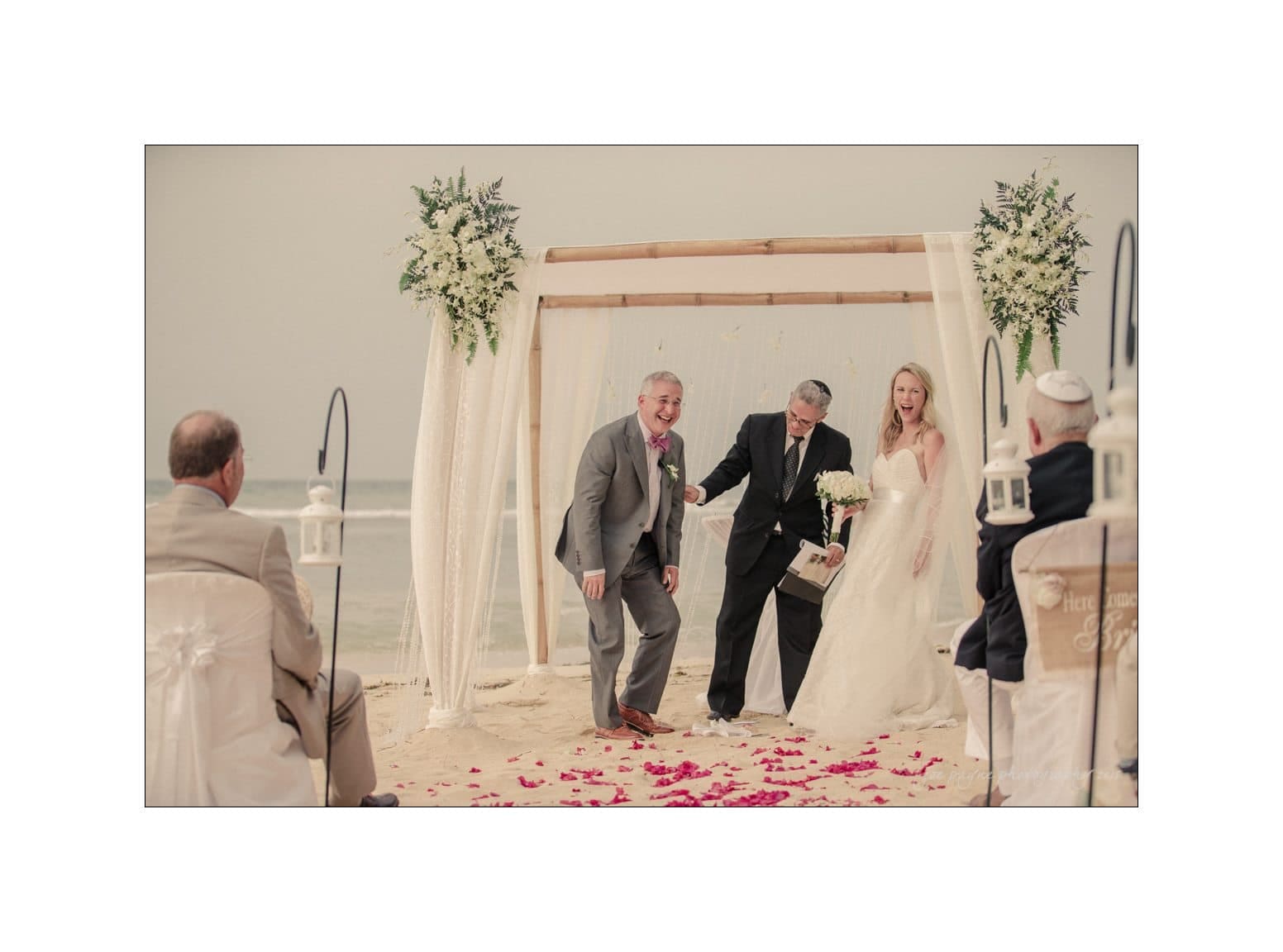 A bride and groom laugh at the beach altar during their wedding ceremony. An officiant stands with them under a canopy adorned with flowers. Guests are seated nearby on white chairs, with rose petals scattered on the sand.
