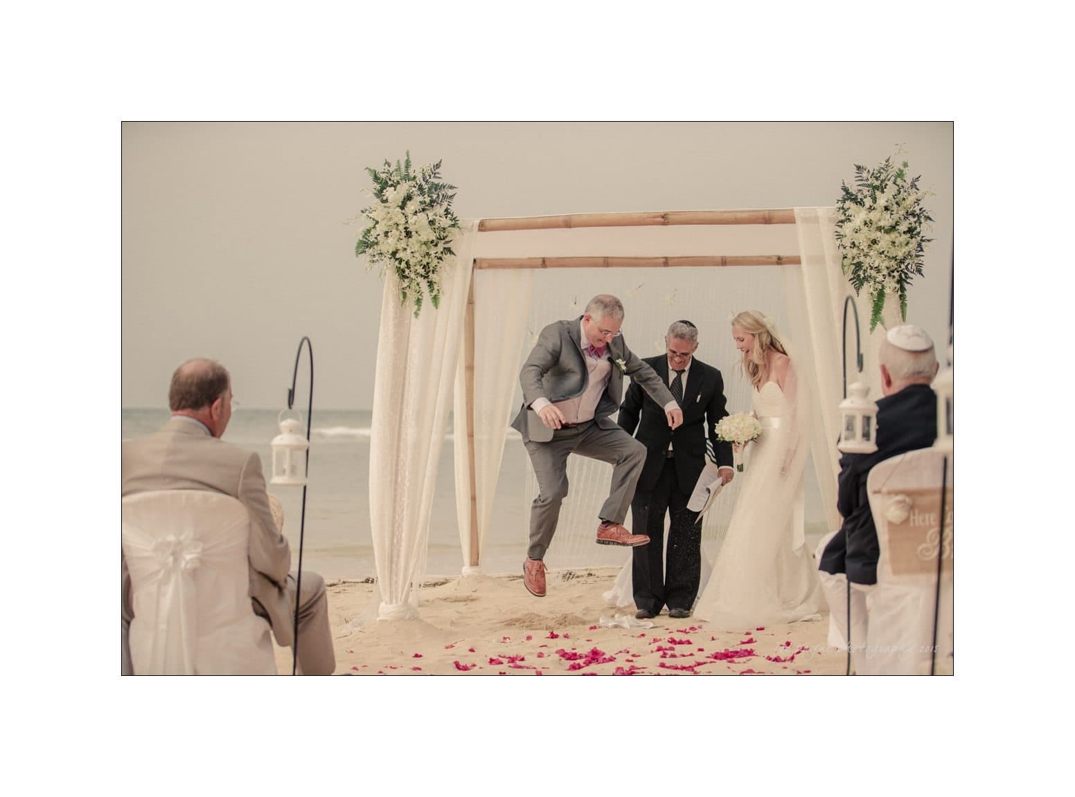 A beach wedding ceremony under a floral canopy. A bride and groom stand facing each other as the groom, in a suit, steps on a glass, following a tradition. Guests are seated, witnessing the event on the sand.