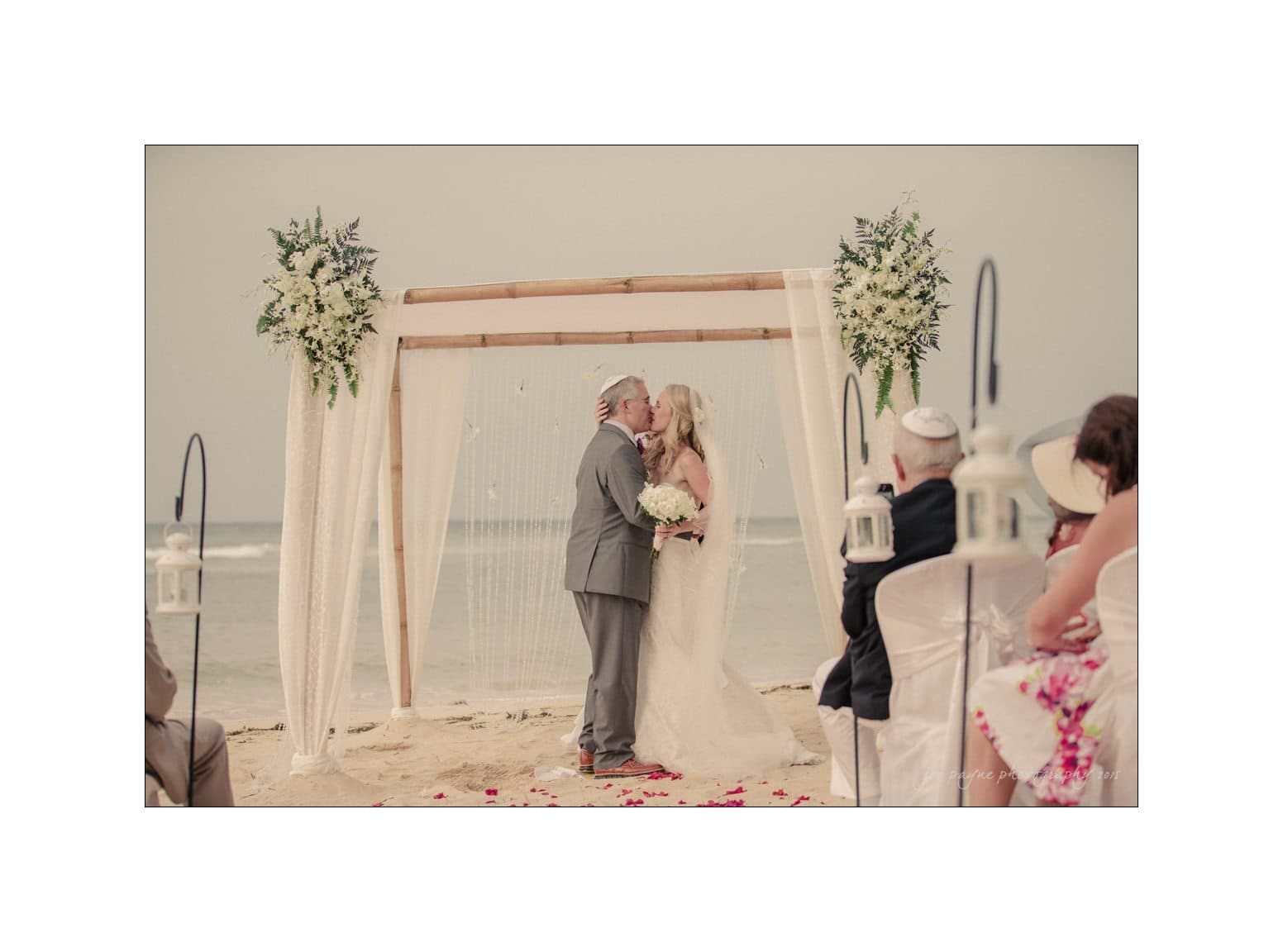 A couple kisses under a floral arch during a beach wedding ceremony. Guests are seated in white chairs, witnessing the moment. The ocean and sky create a serene background.
