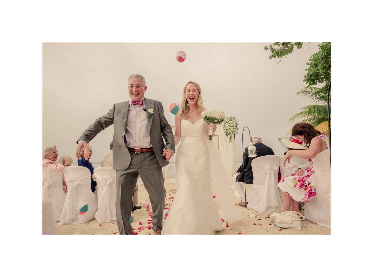 A joyful bride and groom walk down an aisle on a beach. The groom wears a gray suit, and the bride wears a white gown, holding a bouquet. Guests in white chairs are tossing beach balls and smiling under a cloudy sky with scattered rose petals.