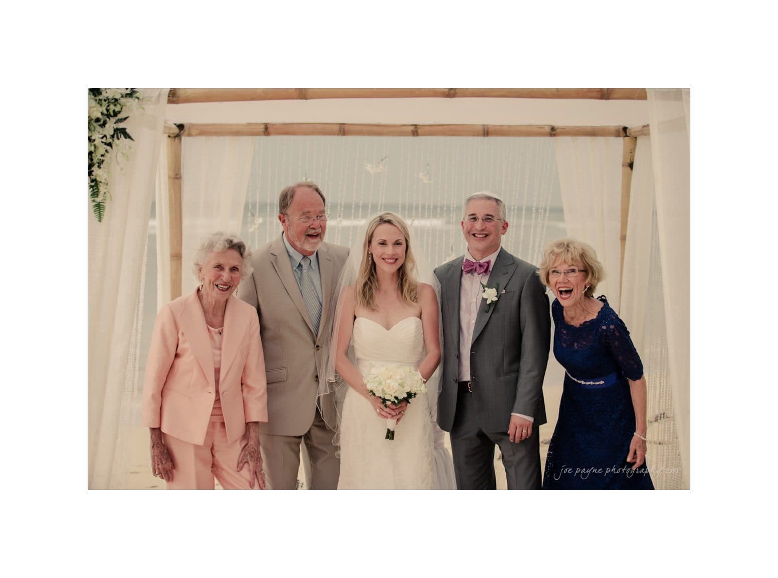 A bride in a white dress holding a bouquet stands under a wooden arch with four older adults, three wearing suits and one in a blue dress. They are all smiling at the camera, with a beach background behind them.