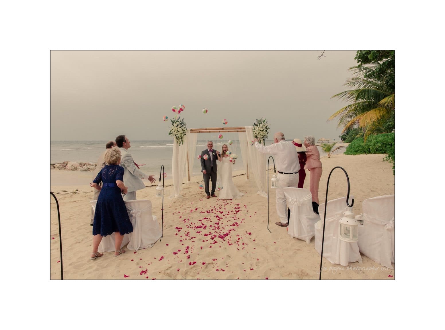 A couple walks down a sandy beach aisle adorned with rose petals after their wedding ceremony. Guests cheer and throw flowers in the air. A floral arch and the ocean create a picturesque backdrop.