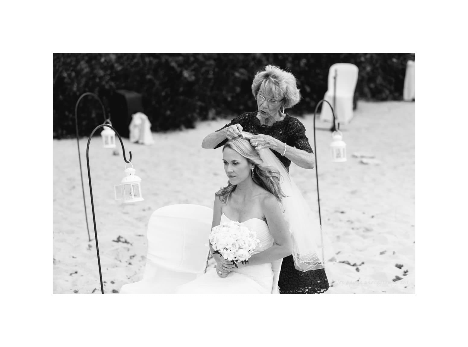 A bride holding a bouquet sits on a sandy beach as an older woman adjusts her veil. The scene is surrounded by decorative lanterns.